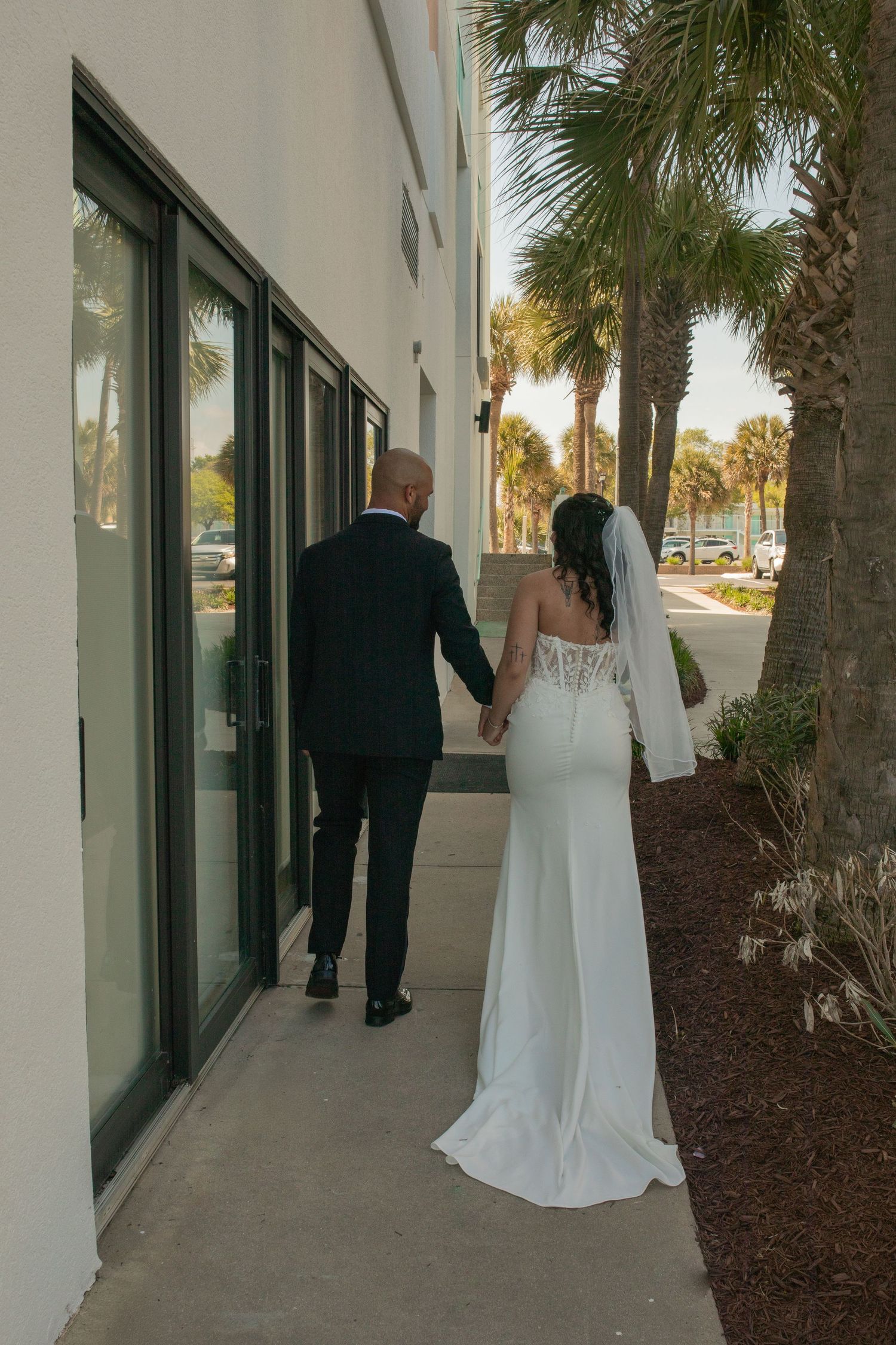 Bride and groom holding hands walking along a modern building exterior with palm trees at their wedding venue.