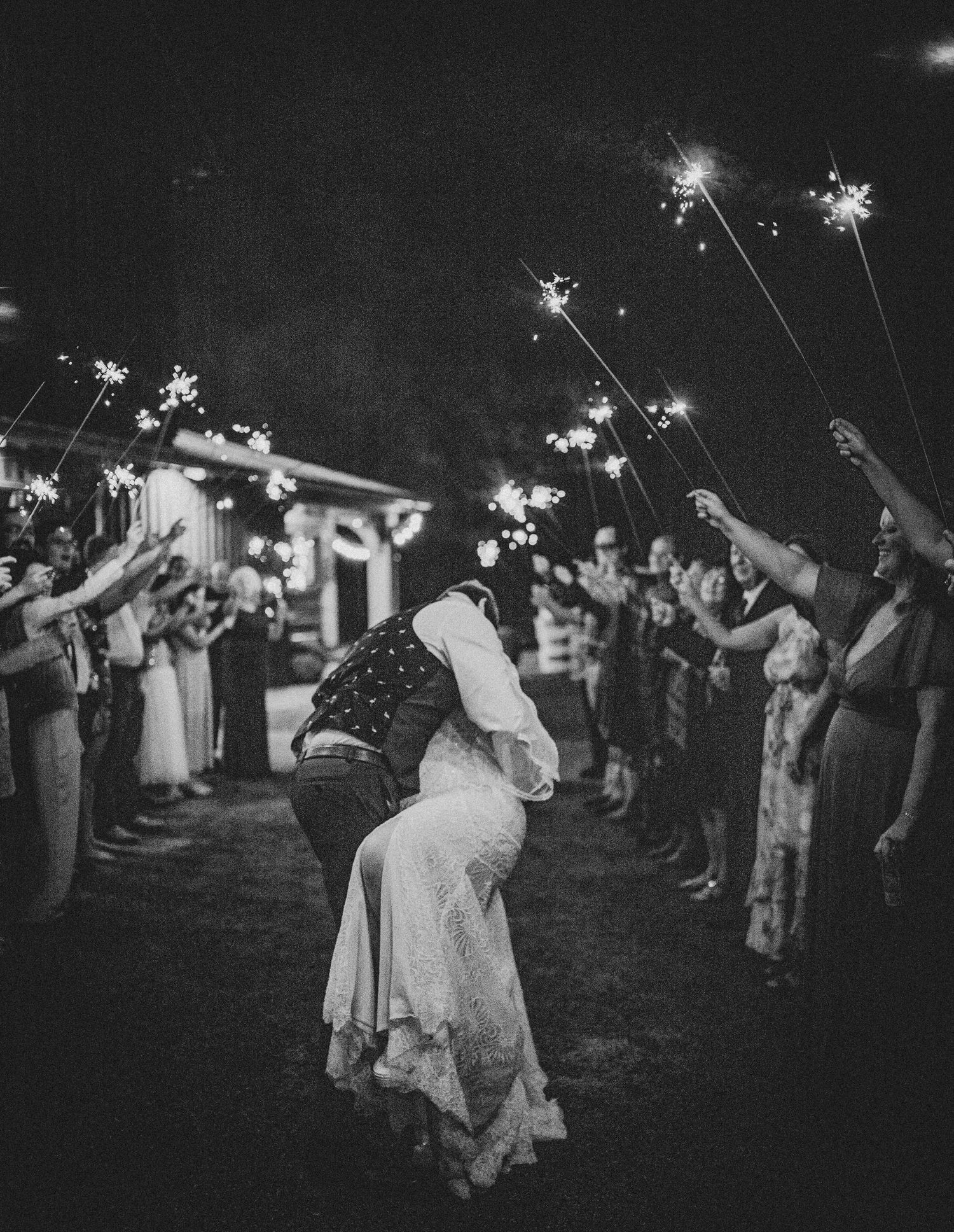 A romantic dip kiss captured in black and white as sparklers illuminate a nighttime wedding celebration.