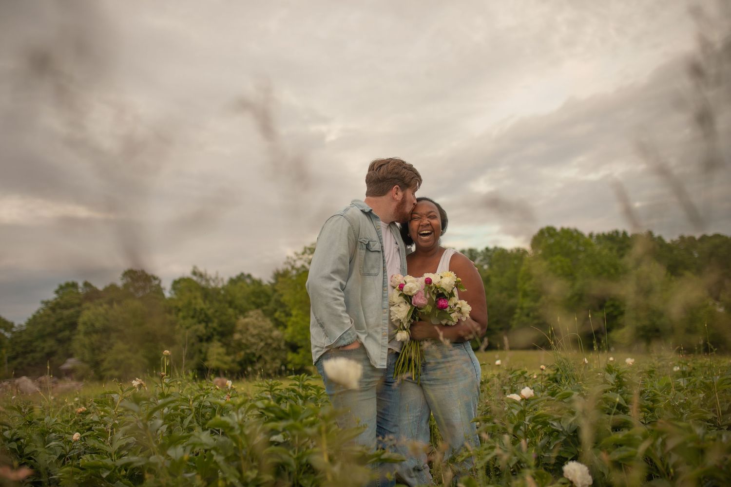Couple sharing a romantic moment in a flower field, man kissing woman's cheek as she holds a bouquet under cloudy skies.