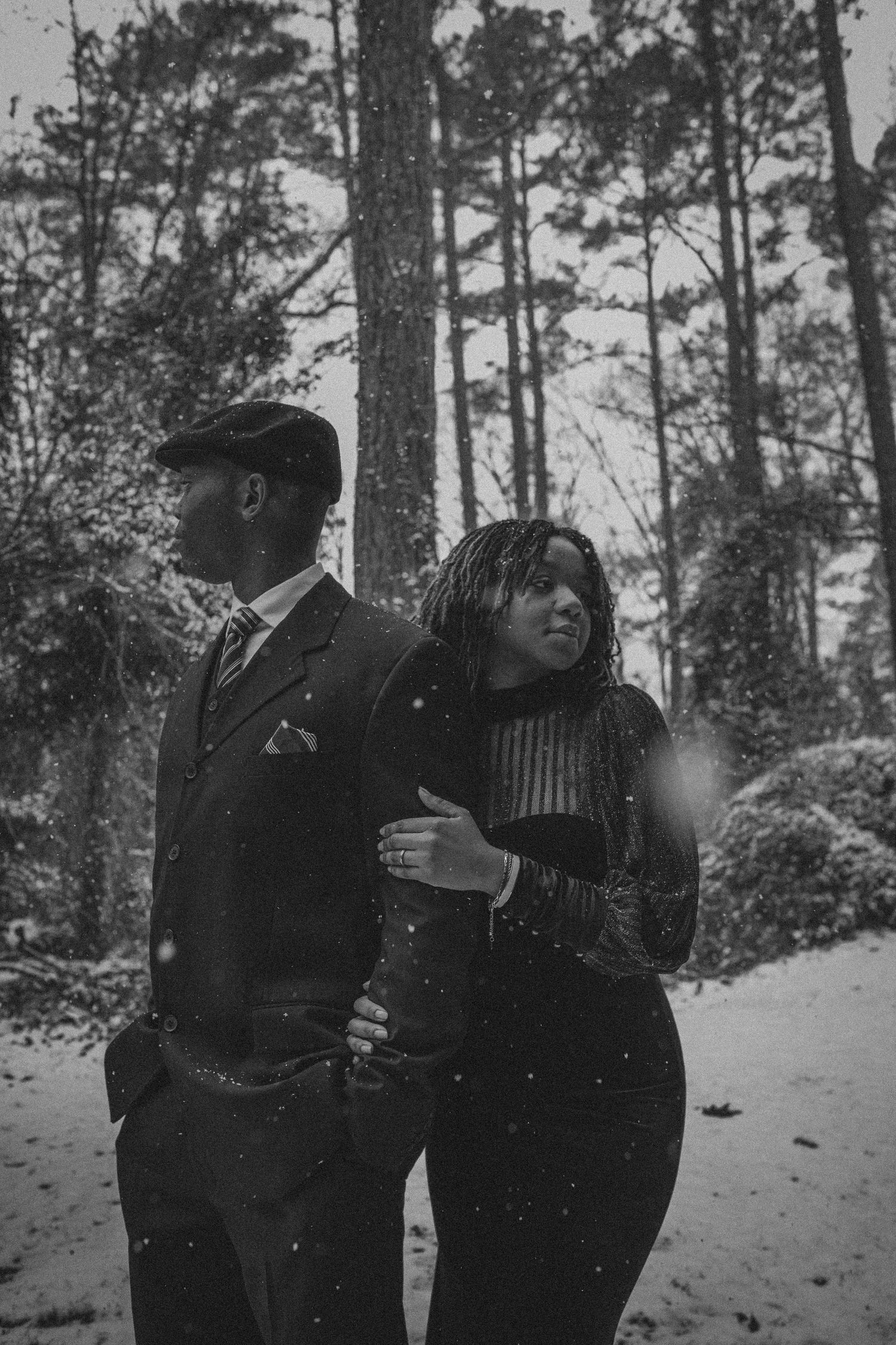 A couple embraces in a snowy forest setting with tall pine trees in the background during winter.