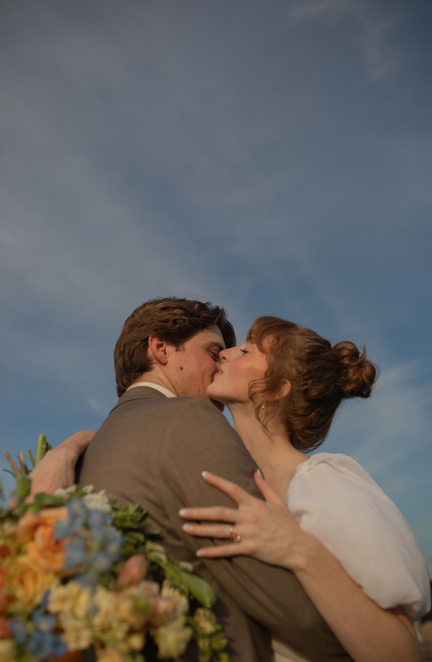 A bride and groom share a romantic kiss against a blue sky backdrop, with colorful wedding flowers in the foreground.