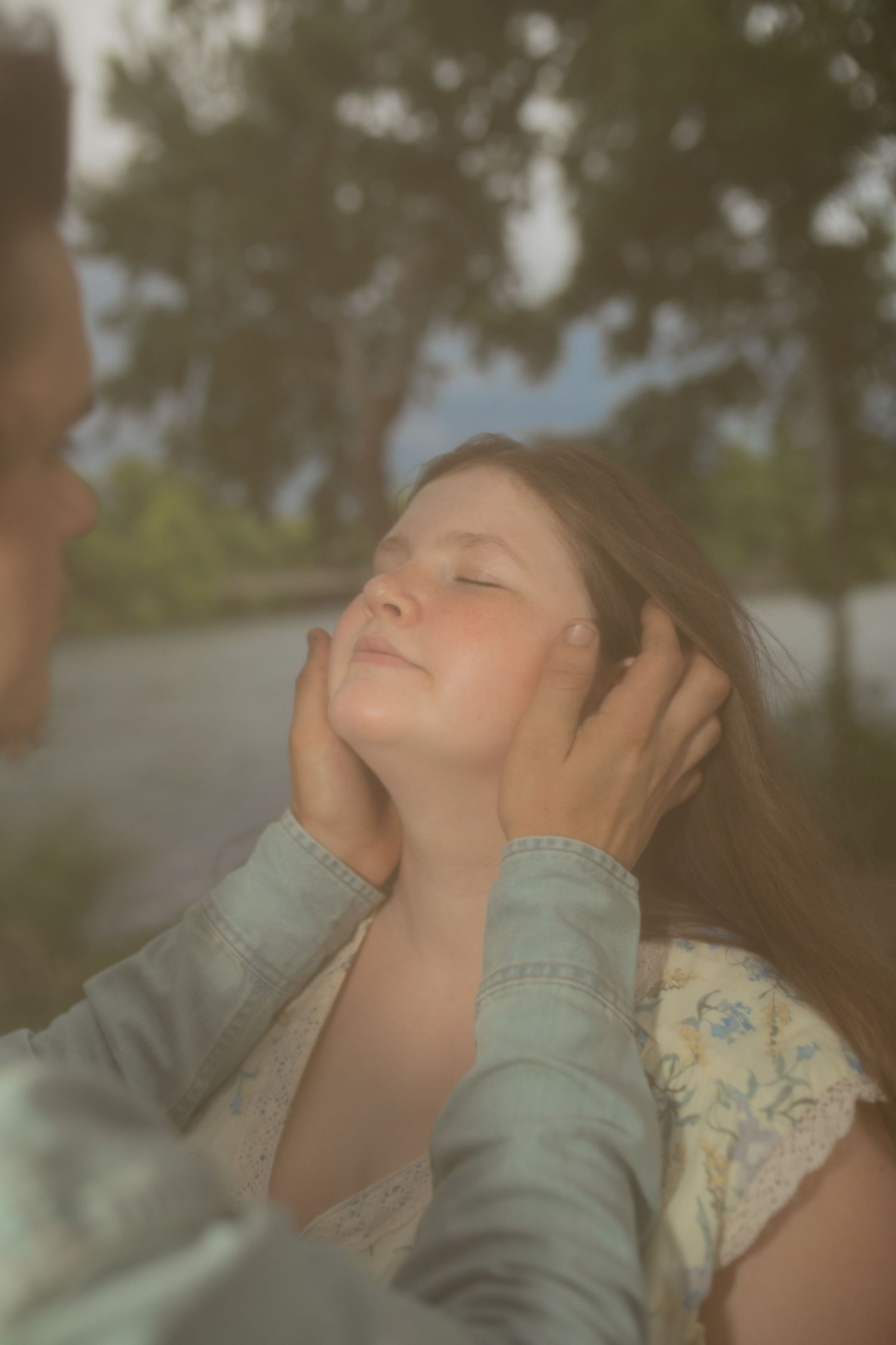 A woman gently cradles a young girl's face with both hands outdoors, as the girl closes her eyes peacefully.