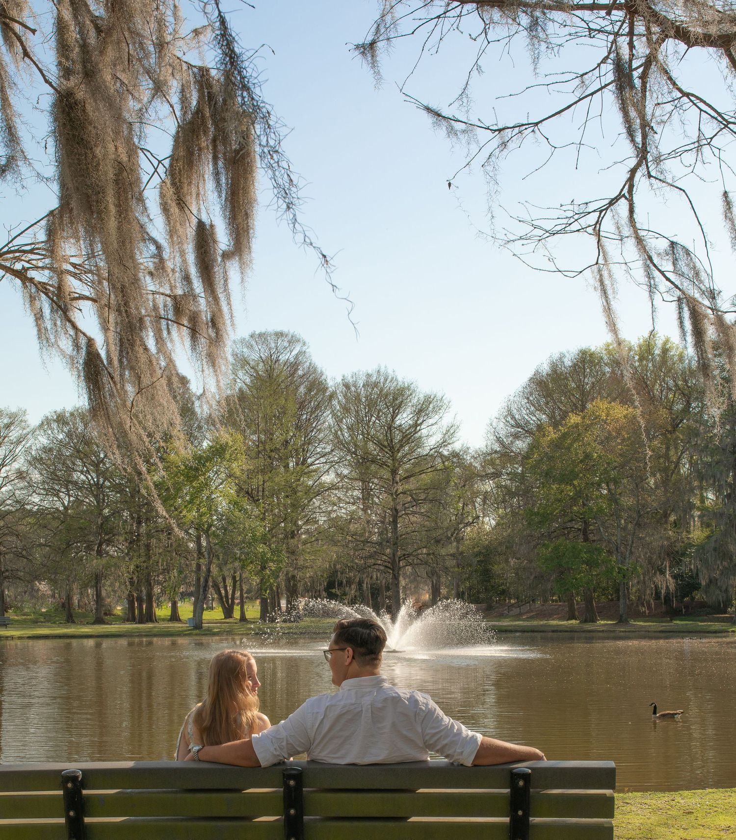 Two people sit on a park bench overlooking a scenic pond with a fountain and Spanish moss hanging from trees.