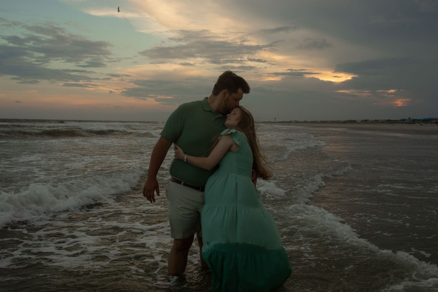 Two people embrace on a beach at sunset as waves wash over the shore and dramatic clouds fill the sky.