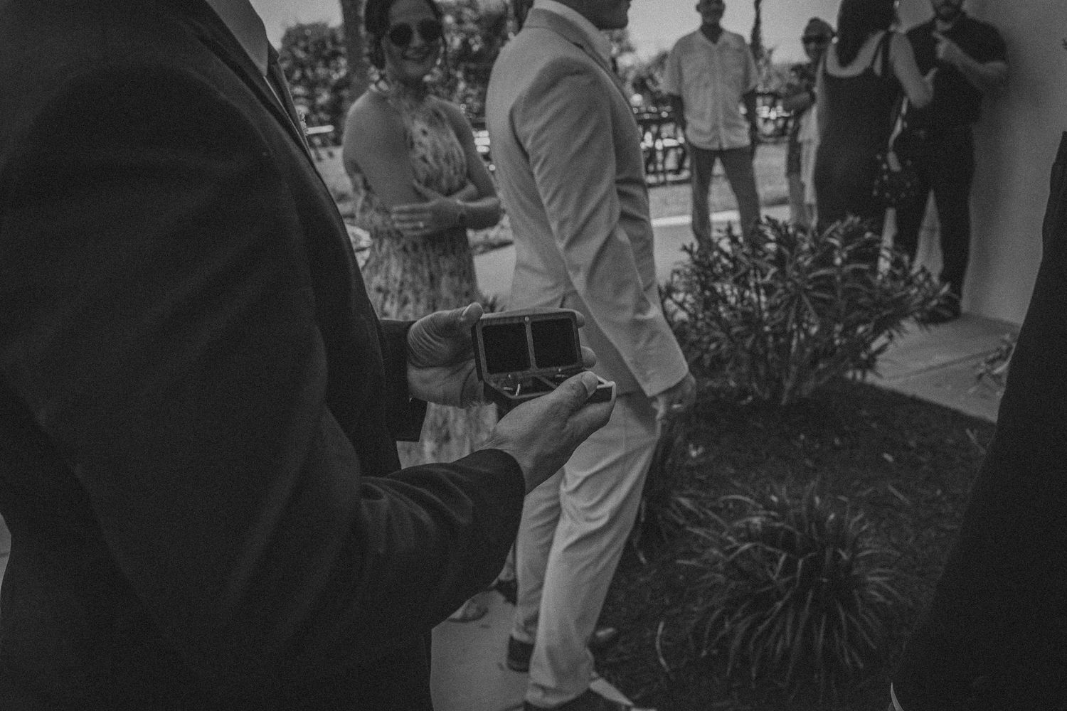 Black and white wedding photo of guests gathered outside, with a man holding a gift box in the foreground.
