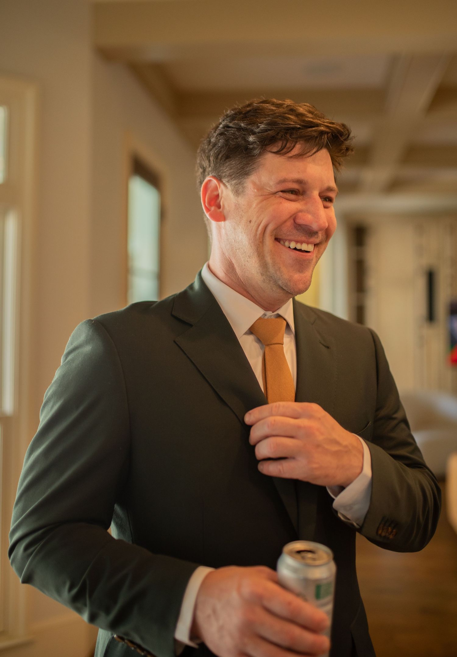 Professional in dark suit and gold tie laughs while adjusting his tie and holding a beverage in an indoor hallway.