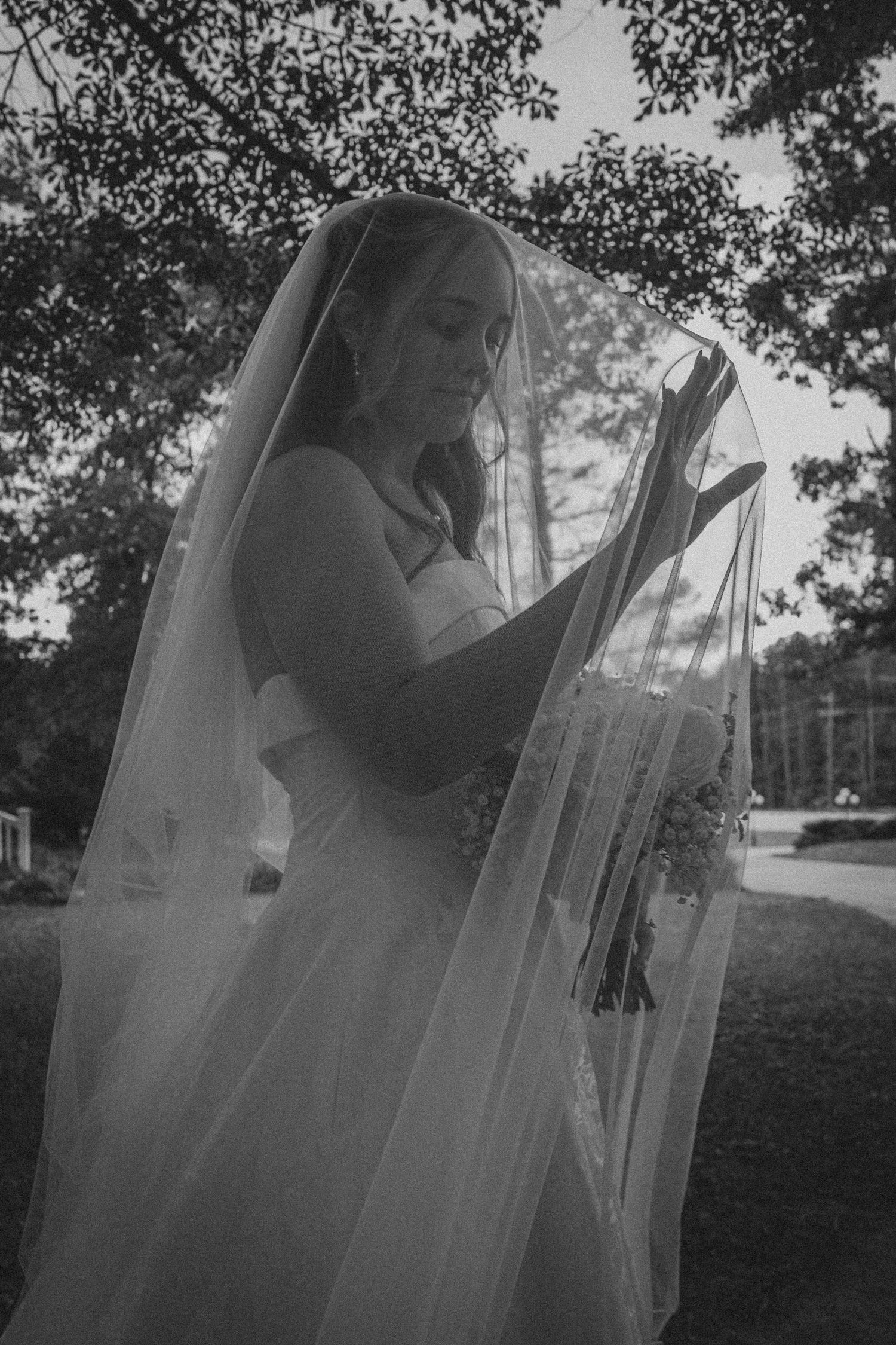 Black and white photo of a bride in a flowing veil holding bouquet outdoors under trees.