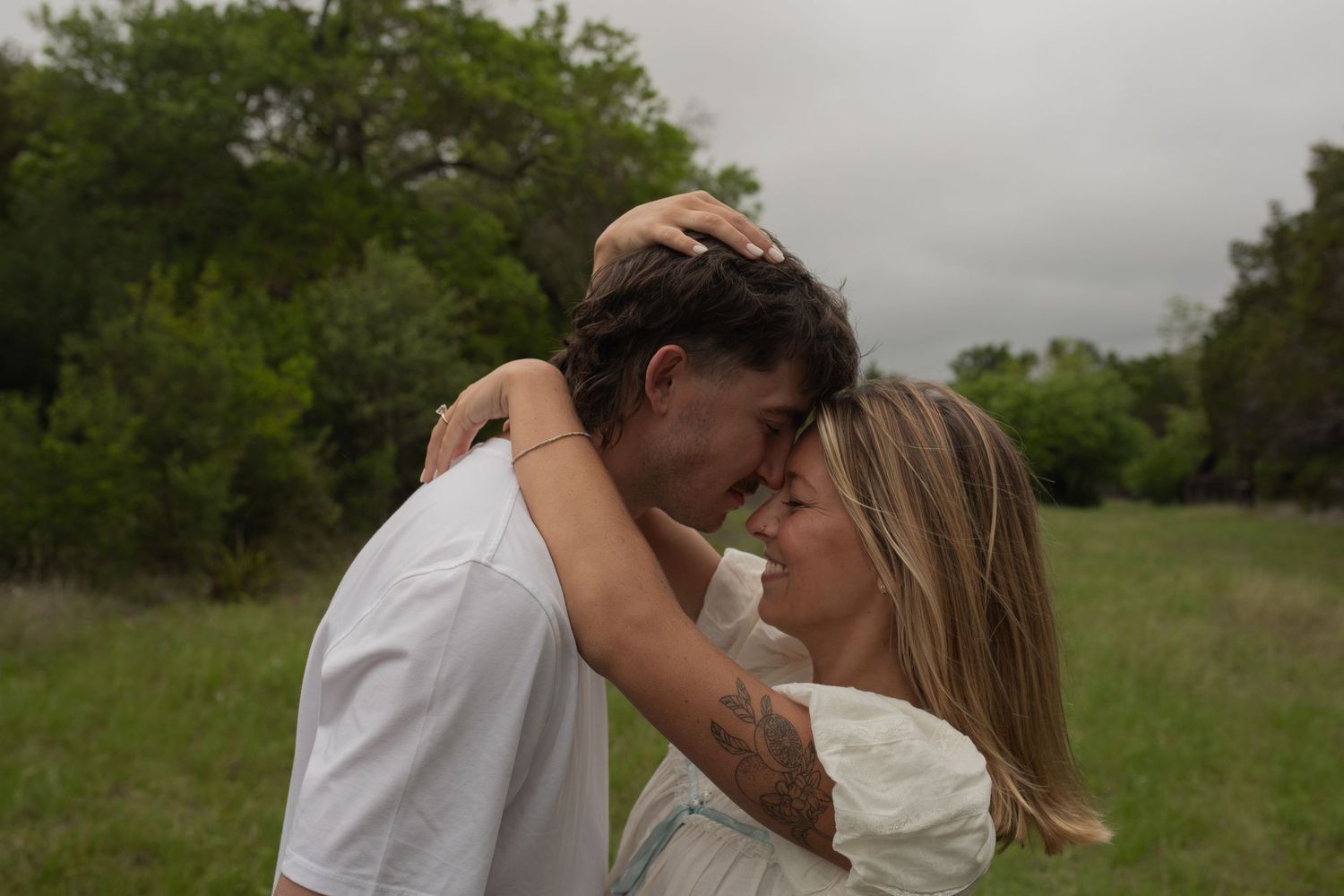 Romantic couple embracing in a green meadow, foreheads touching tenderly, surrounded by lush trees on a cloudy day.