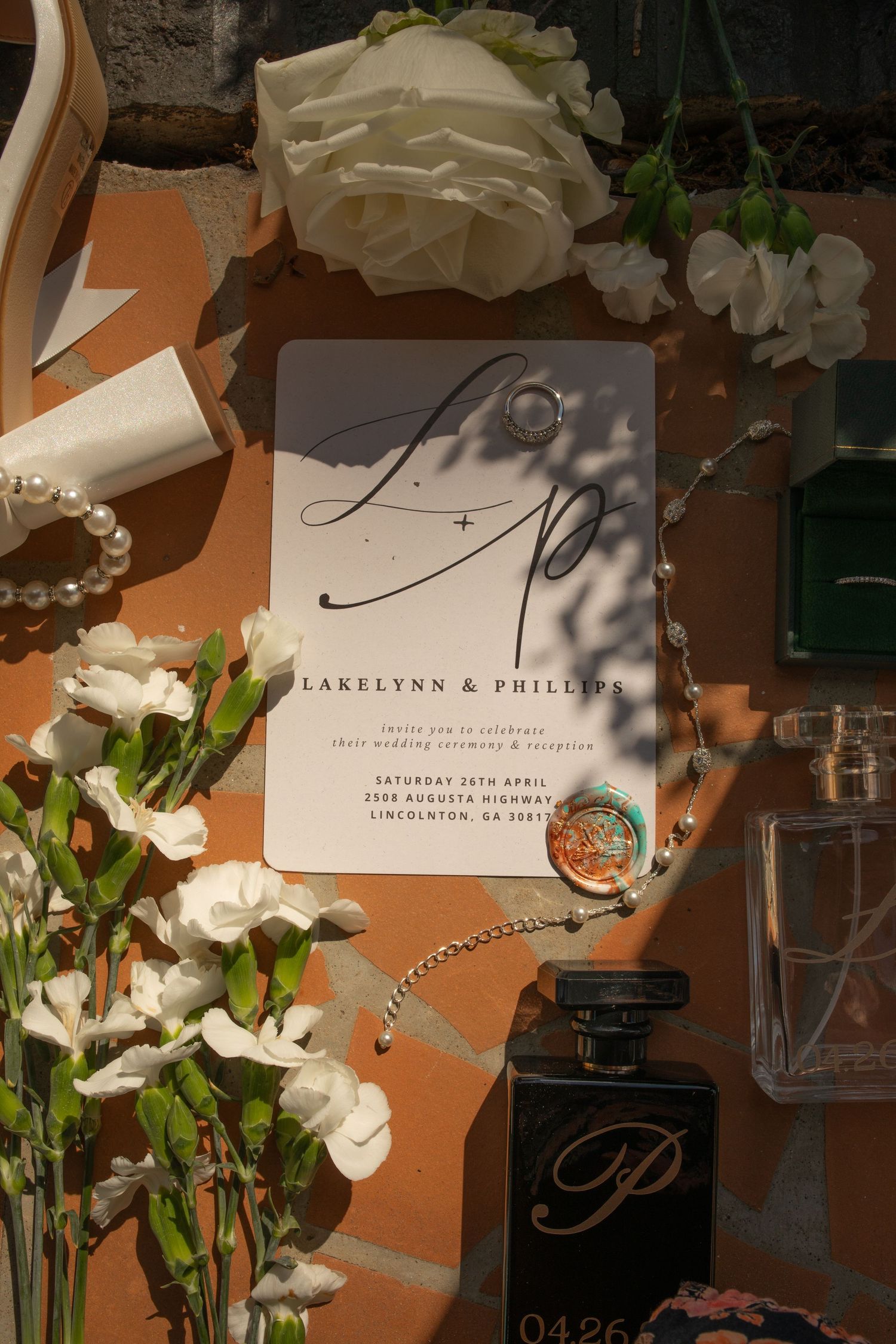 Elegant wedding invitation for Lakelynn & Phillips surrounded by white flowers, pearls, perfume, and jewelry on orange surface.