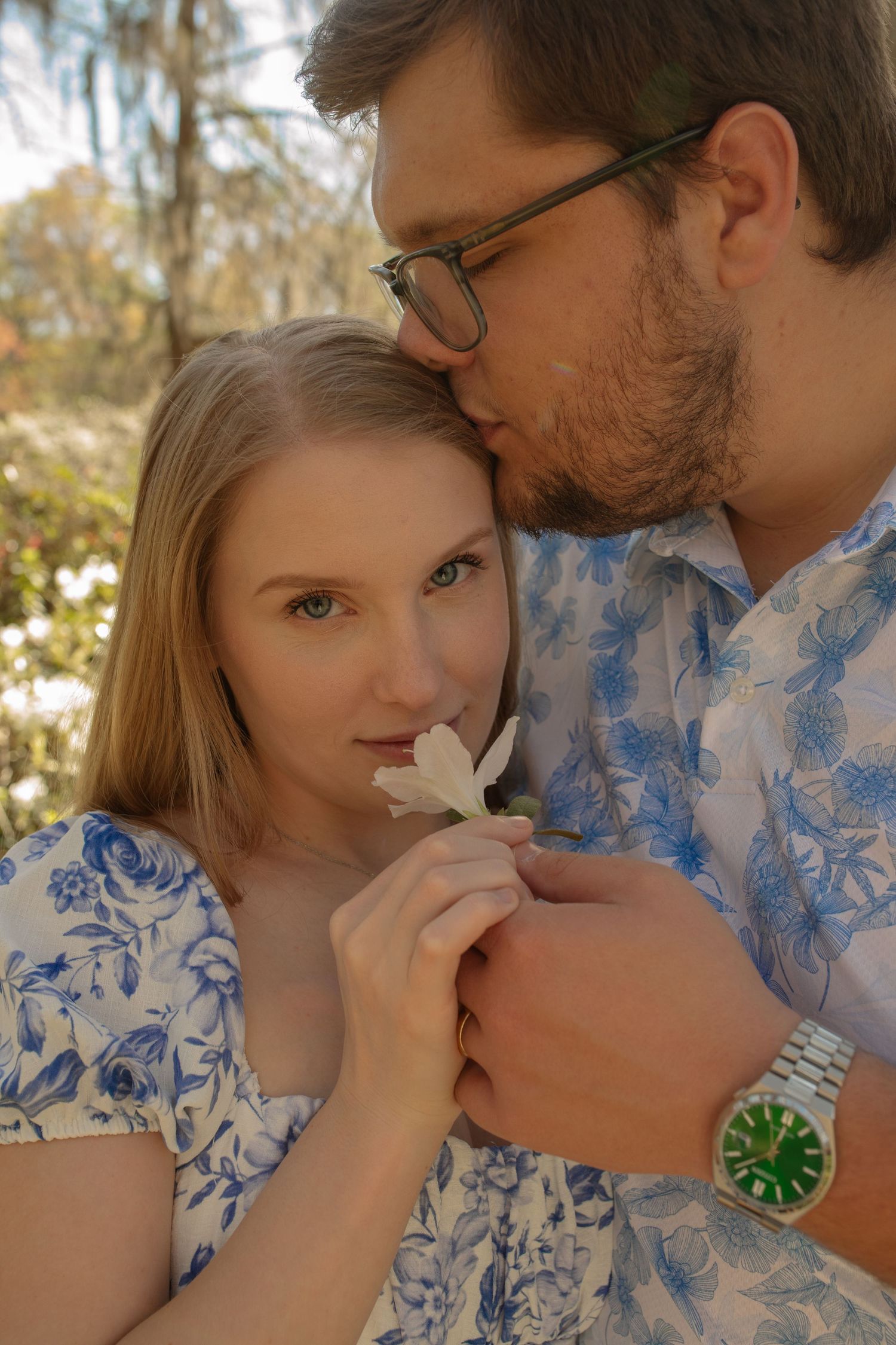 A young couple in matching floral outfits share a tender moment, as the man kisses her head while she holds a white flower.