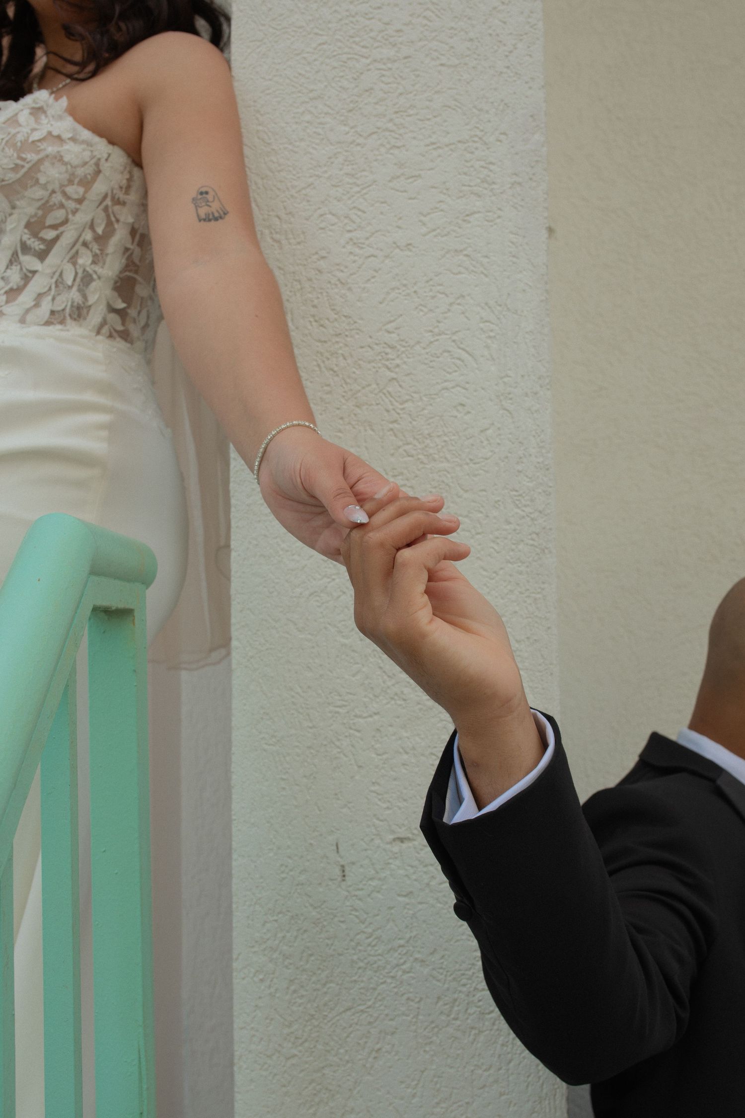 Groom in black suit holds bride's hand with bracelet and tattoo, standing by mint green railing in wedding attire.