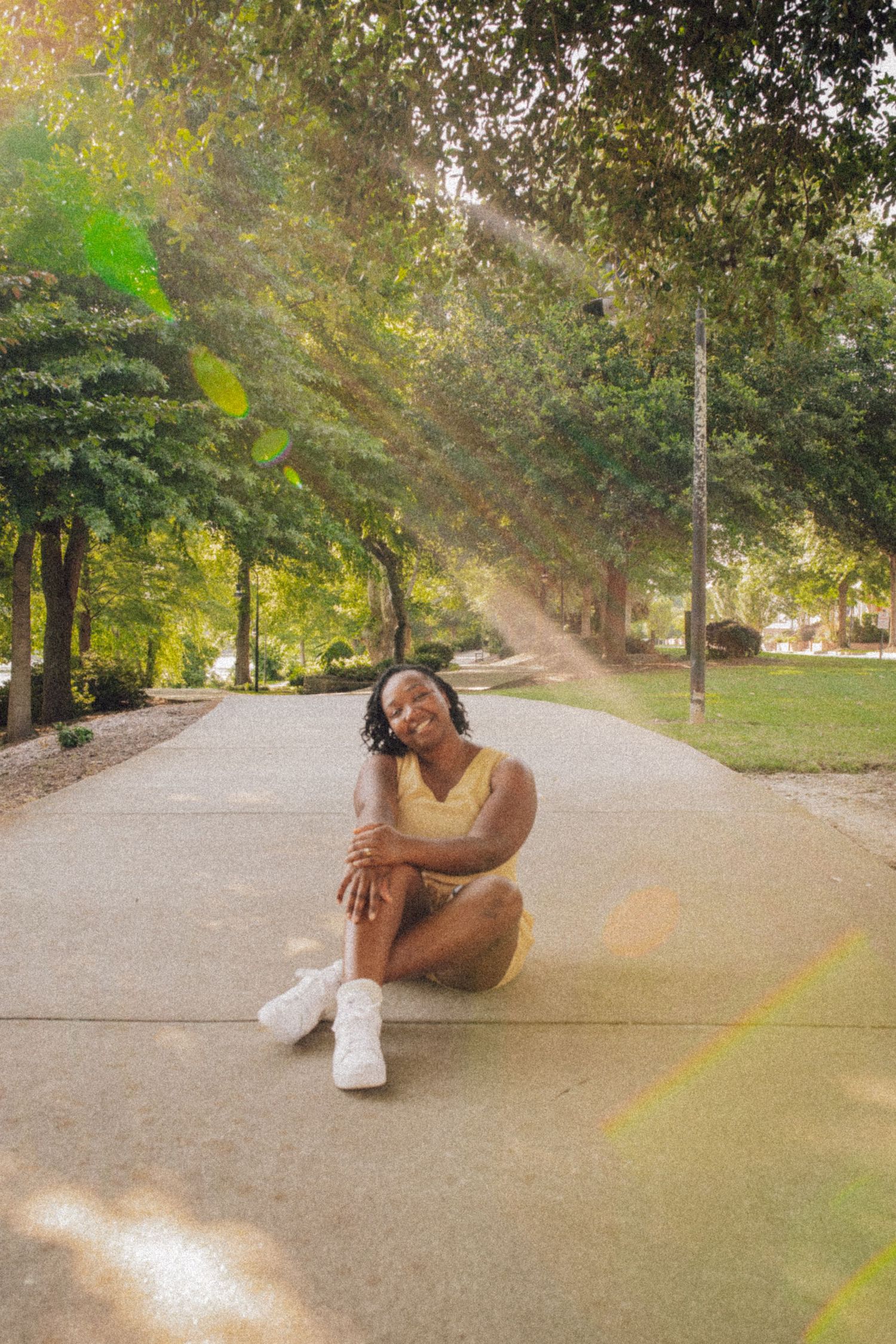 Person in a nude-colored outfit sits on a sunlit pathway surrounded by green trees and foliage on a warm day.