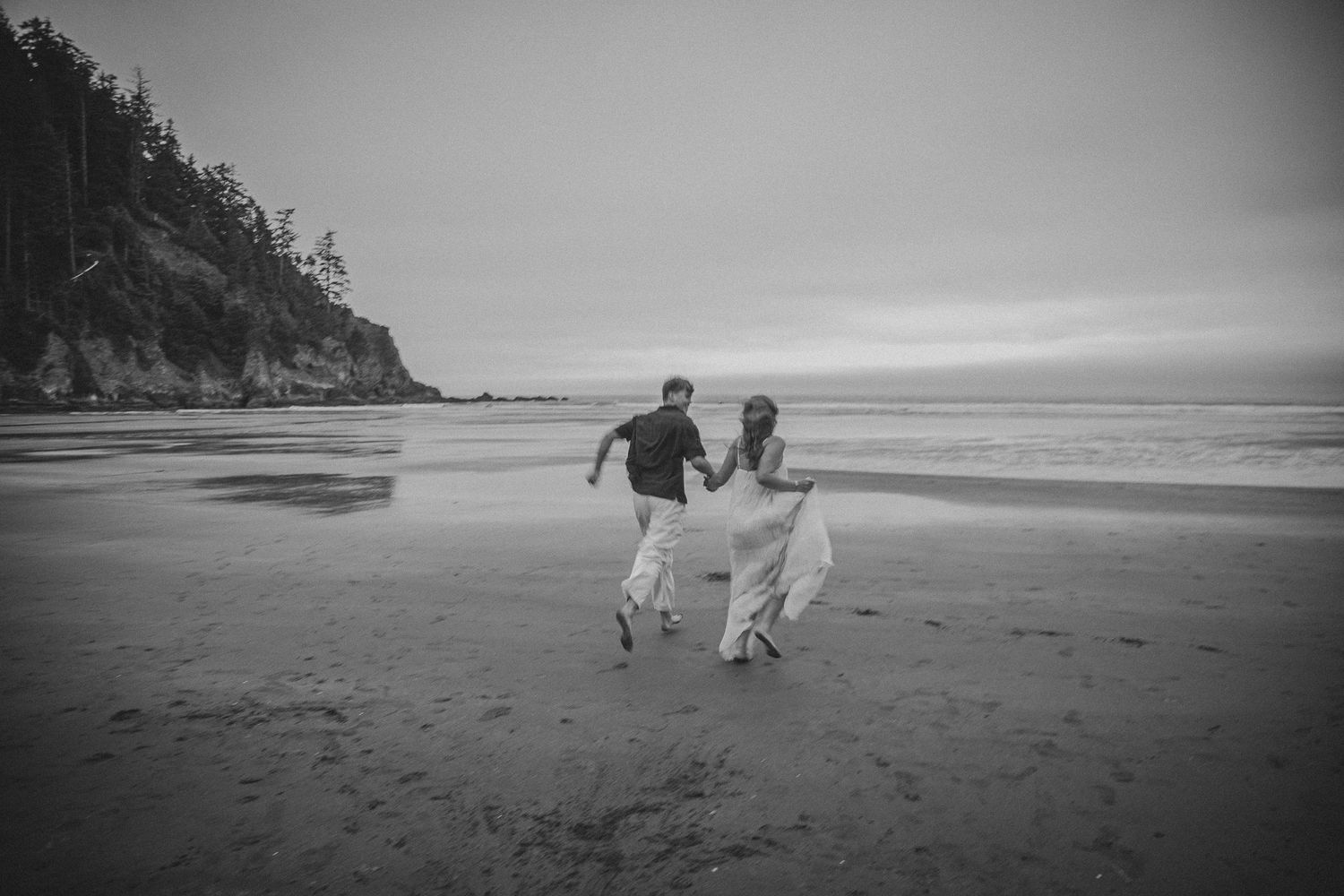 A couple in wedding attire runs joyfully hand-in-hand along a scenic black and white beach shoreline.
