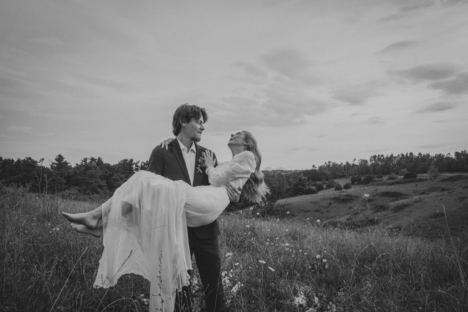 Groom carrying bride in white dress through a meadow in black and white wedding photography outdoors.