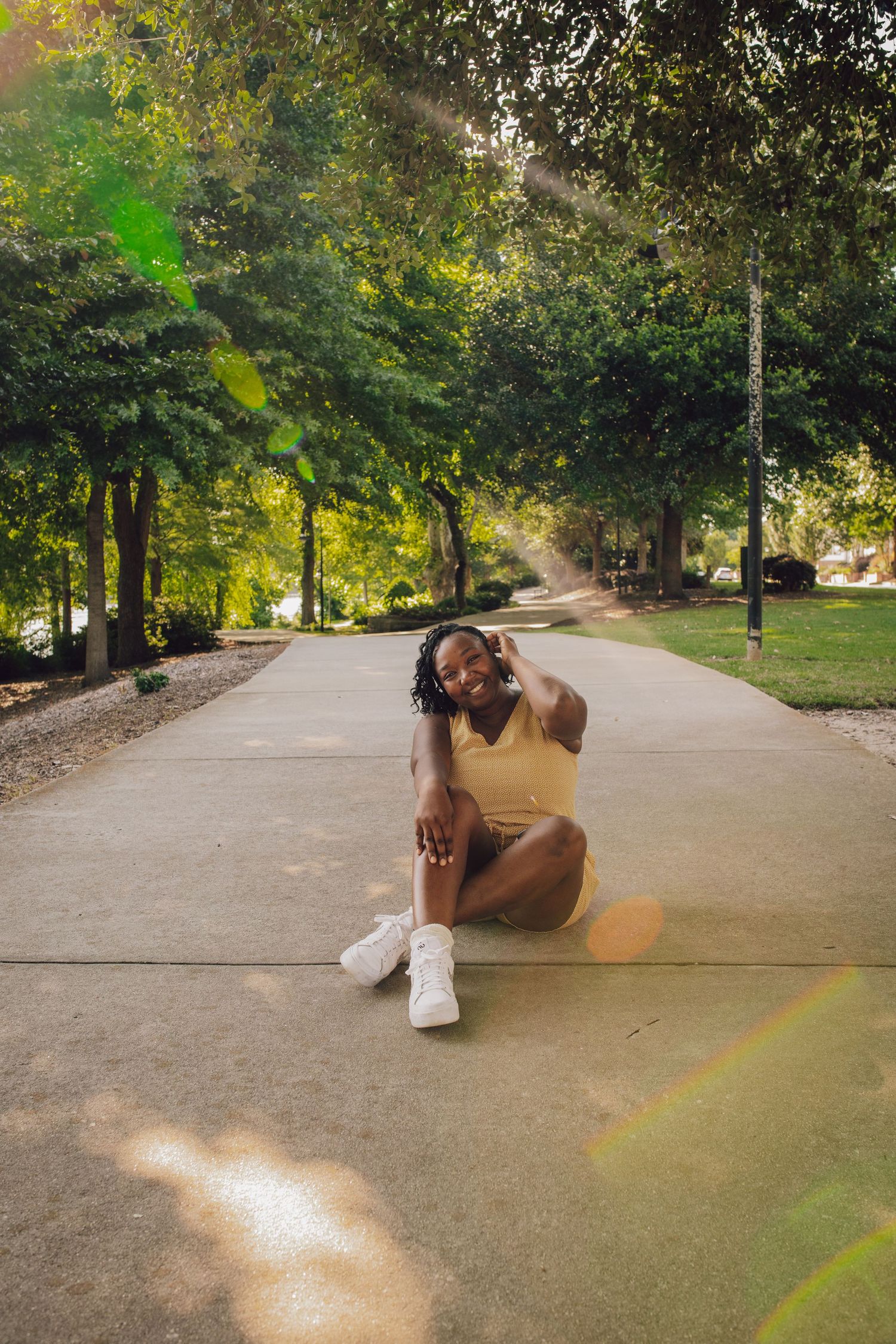 Young Black woman in yellow outfit sitting cross-legged on a sunlit park path, smiling and posing among lush green trees.