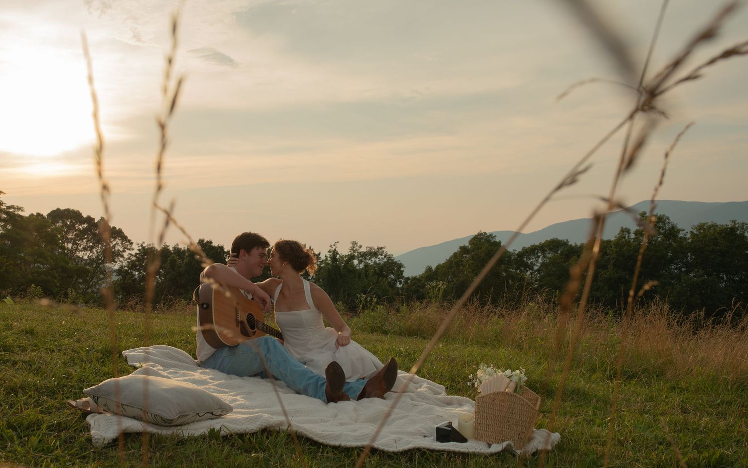 Couple sharing a romantic kiss on a blanket during a golden sunset picnic in a lush green meadow.