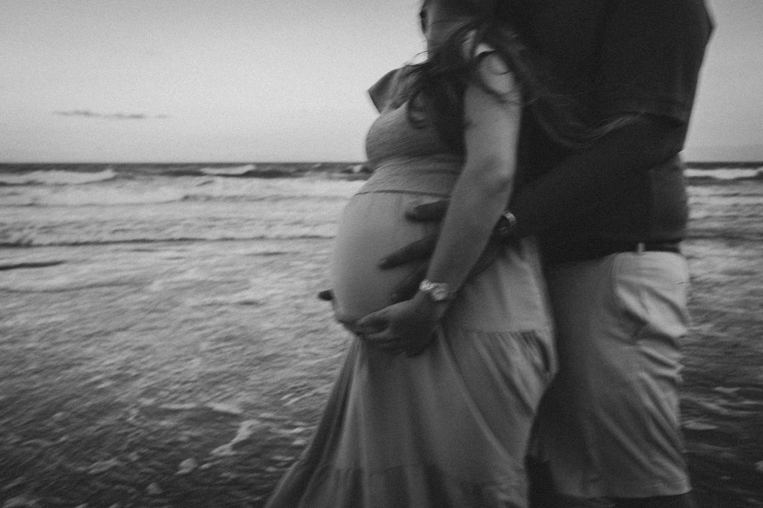 A couple embraces by the ocean during a maternity photo session at sunset, captured in black and white.