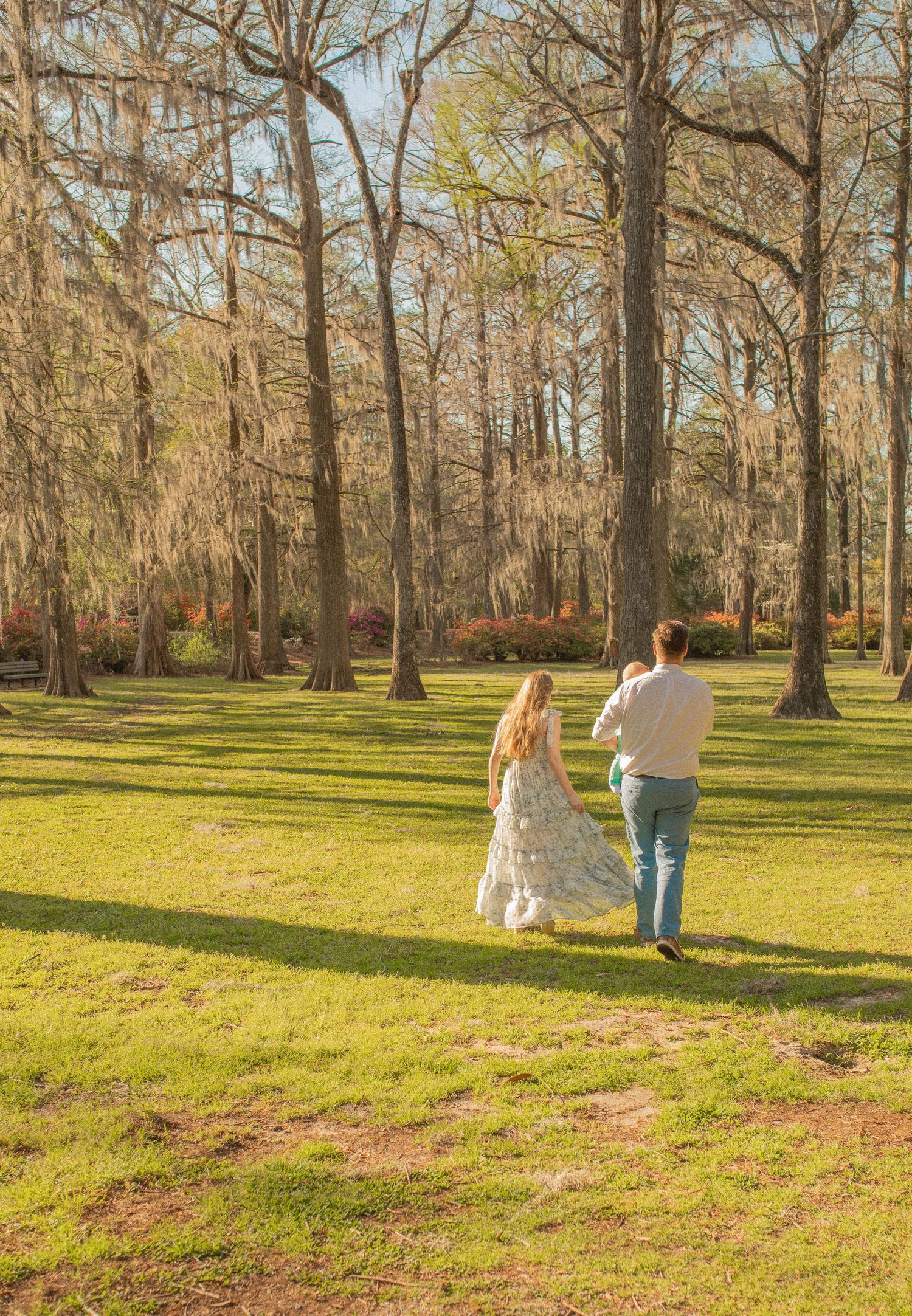 A couple walks together through a sunlit forest park with Spanish moss hanging from tall trees in the late afternoon.