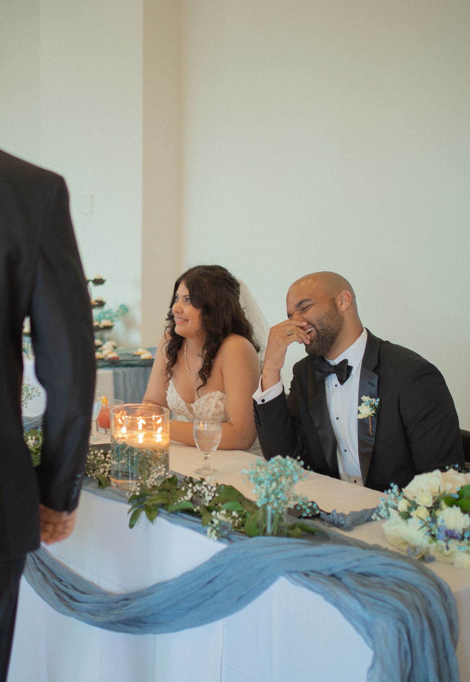 Bride and groom laughing joyfully at sweetheart table decorated with blue draping and candles during wedding reception.
