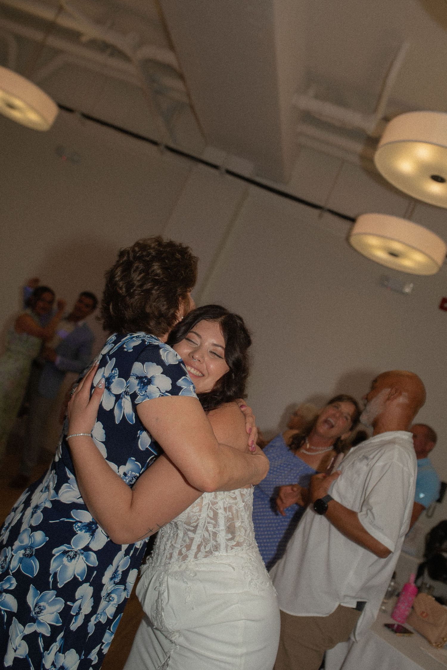 Two women share a warm embrace while dancing at an indoor wedding reception with guests and pendant lights in the background.