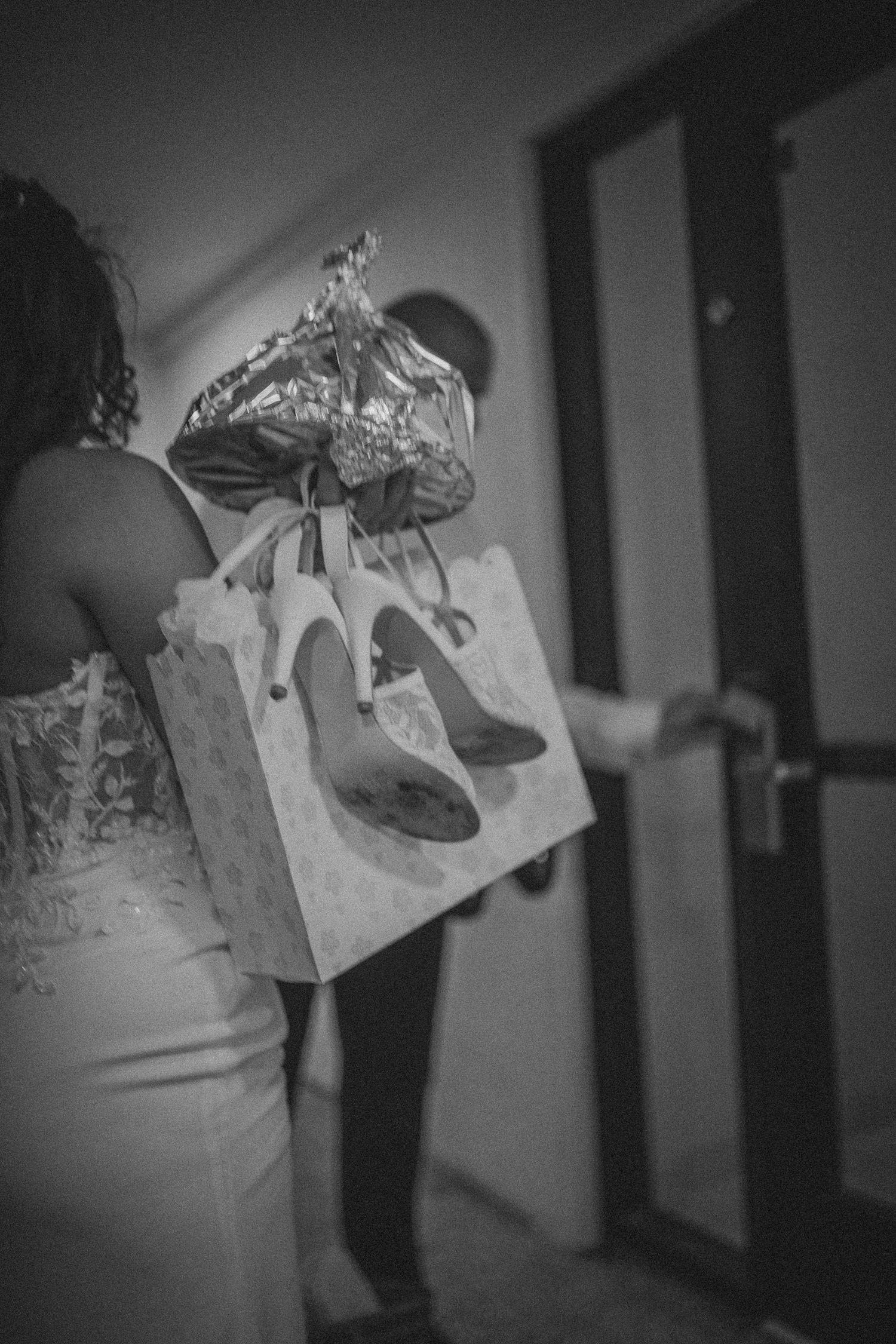Black and white photo of woman in lace dress holding gift bag with balloon and high heel shoes print near door.
