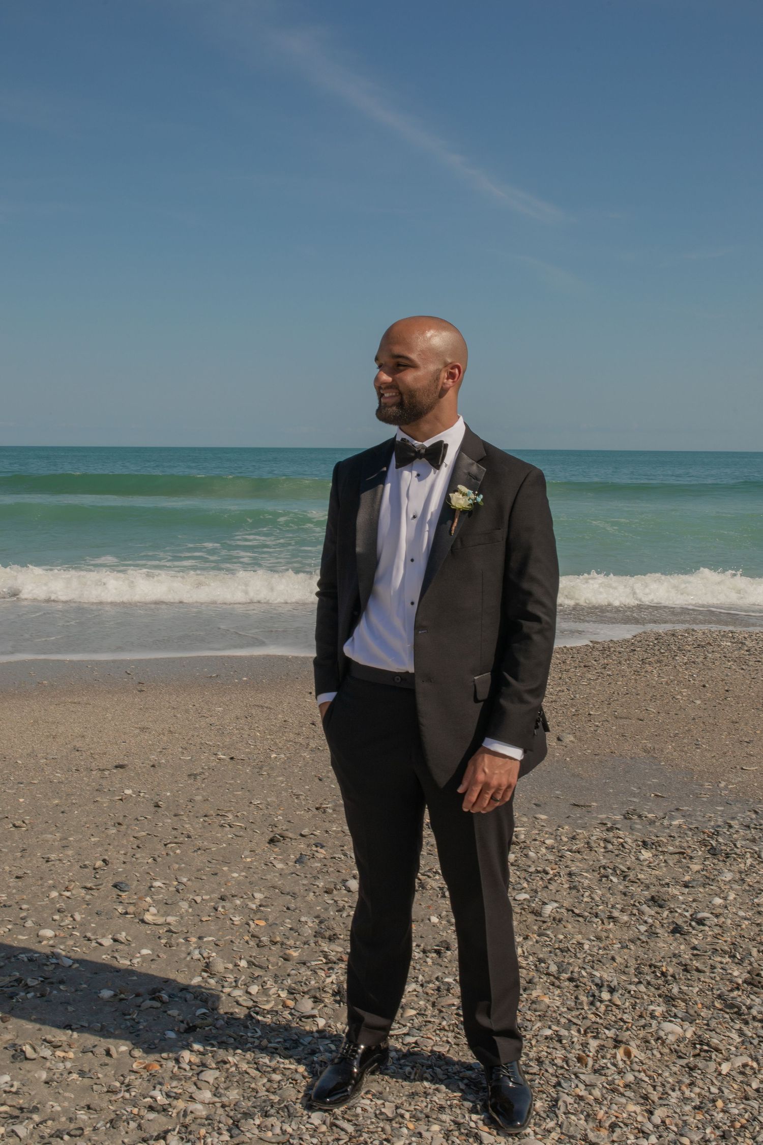 Groom in black tuxedo with boutonniere standing on rocky beach with turquoise ocean waves behind him.
