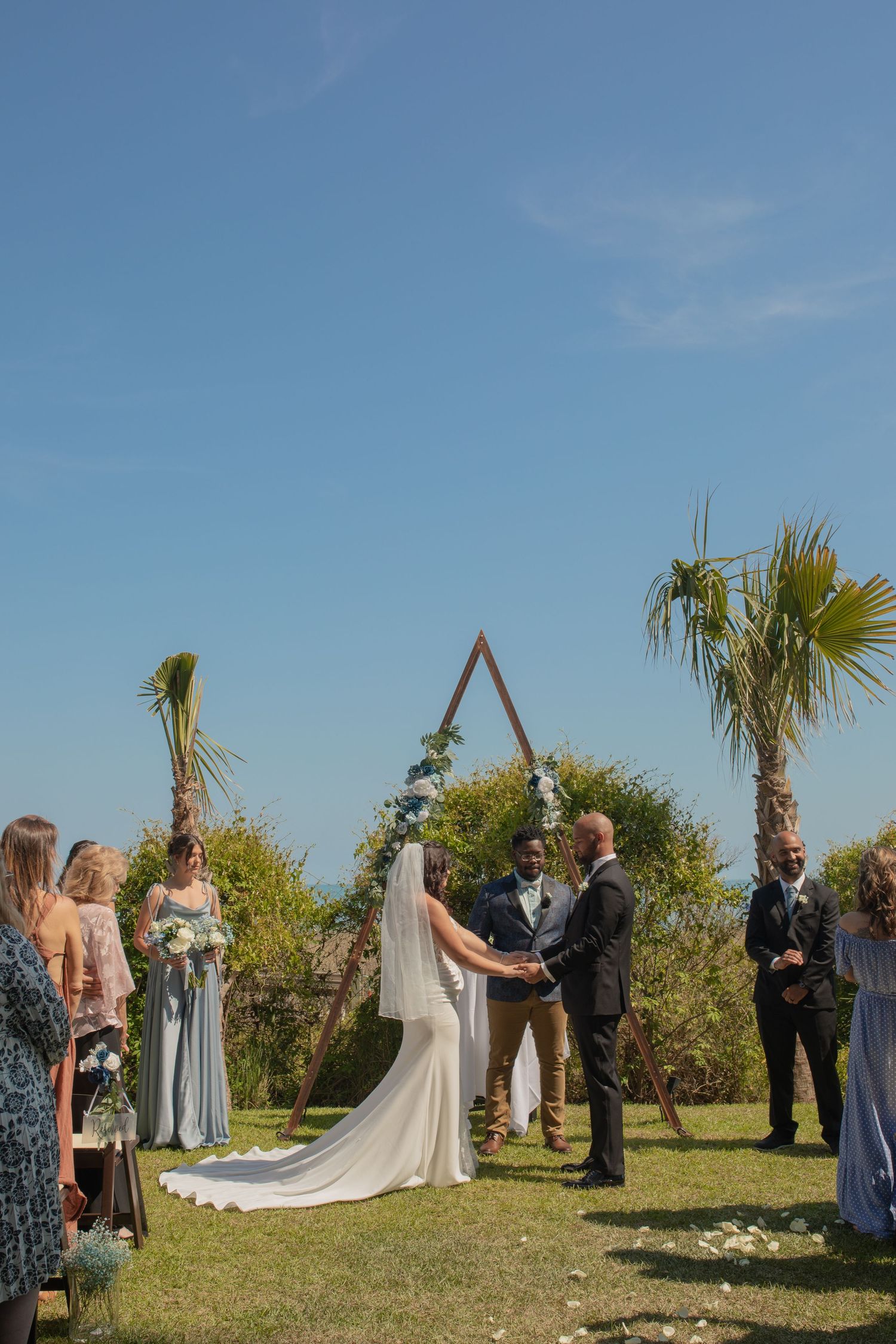 Outdoor wedding ceremony with bride and groom at triangular arch, palm trees, and blue sky backdrop.