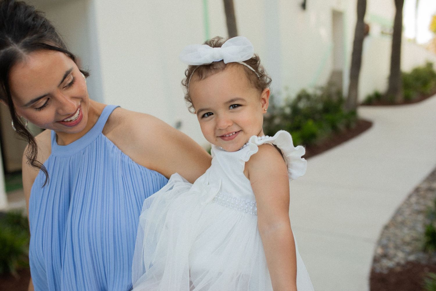 A smiling woman in a blue dress holds a cute toddler girl in a white dress with a bow outdoors.