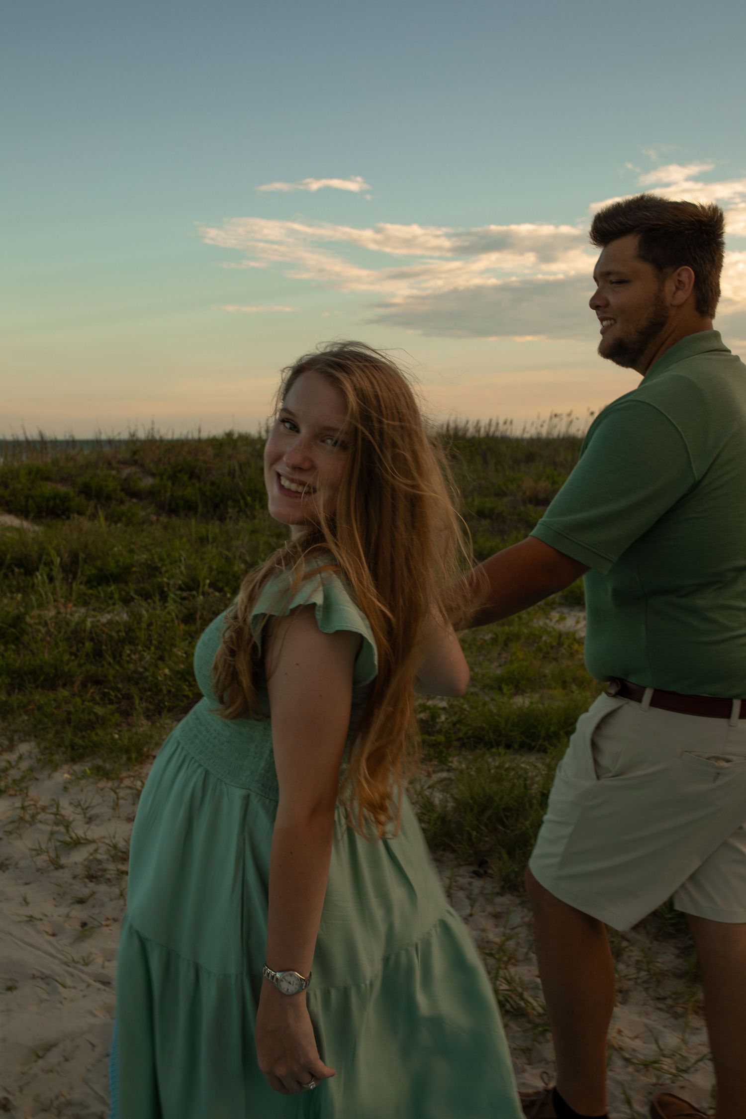 A couple in coordinated outfits enjoys a sunset moment on coastal dunes.