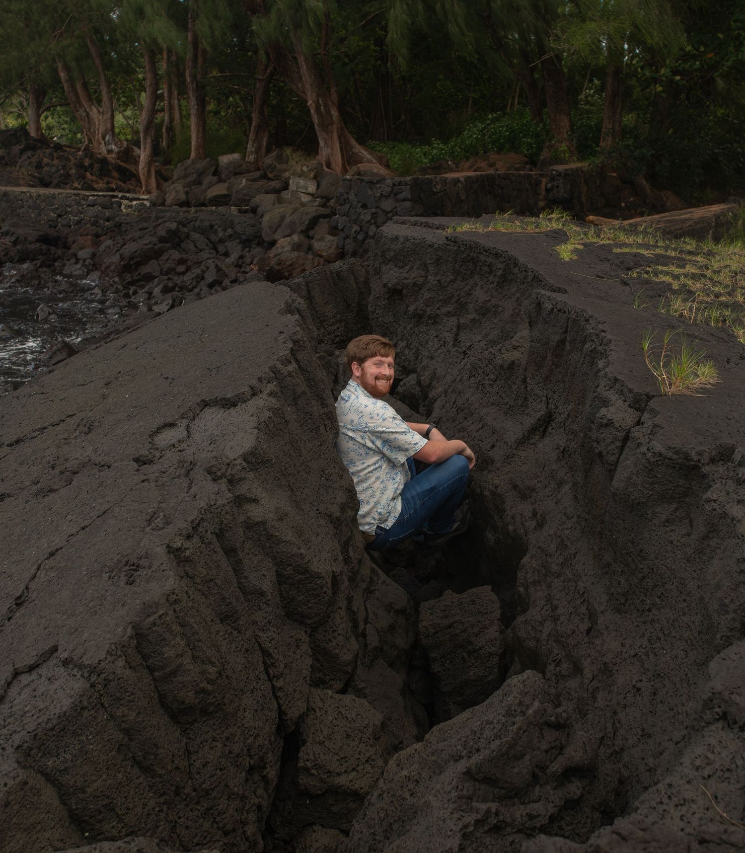 Person sitting on dark volcanic rock formation surrounded by lush tropical forest in the background.