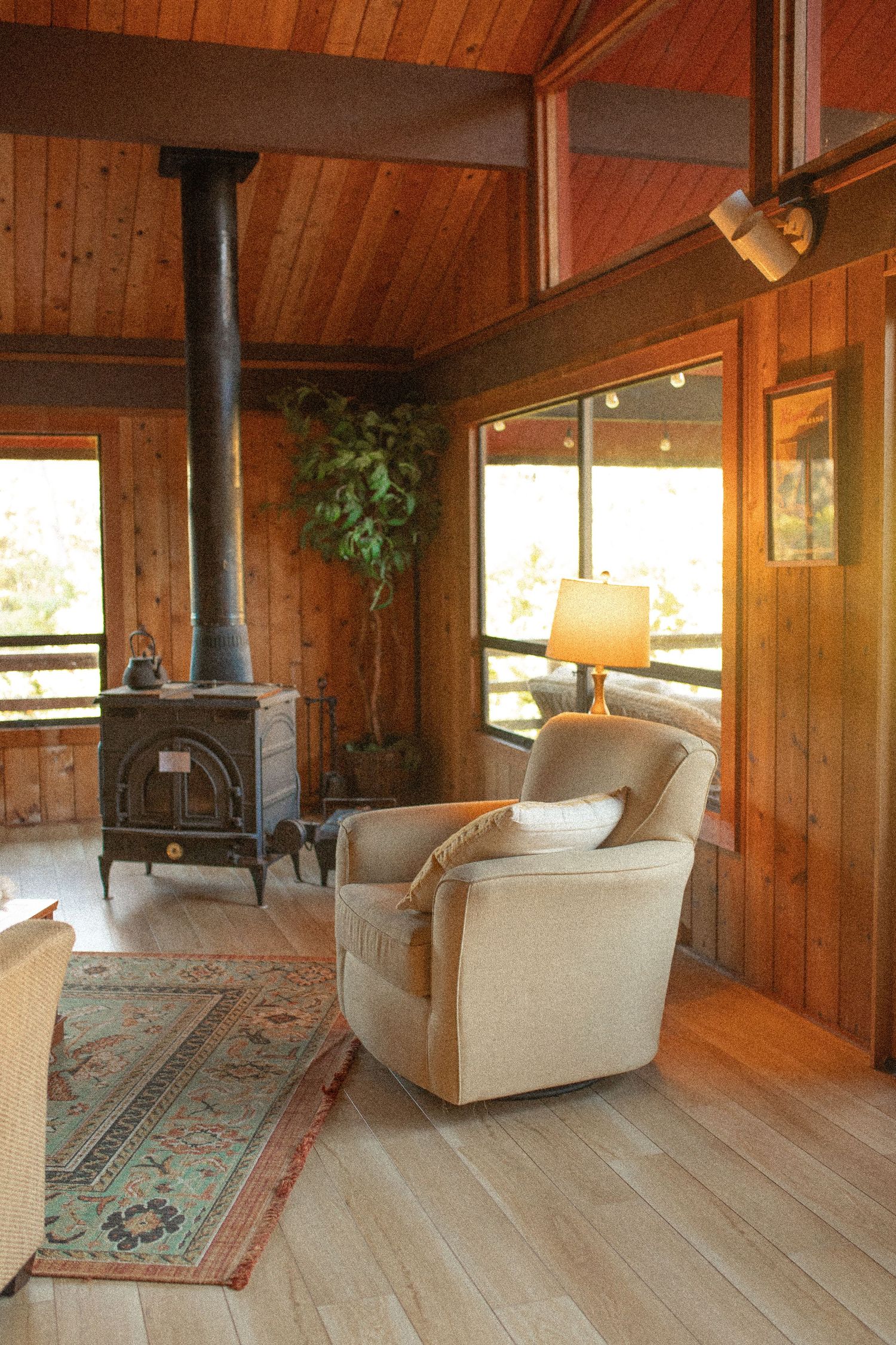 Cozy cabin interior with wood-burning stove, beige armchair, hardwood floors and paneled walls bathed in natural light.