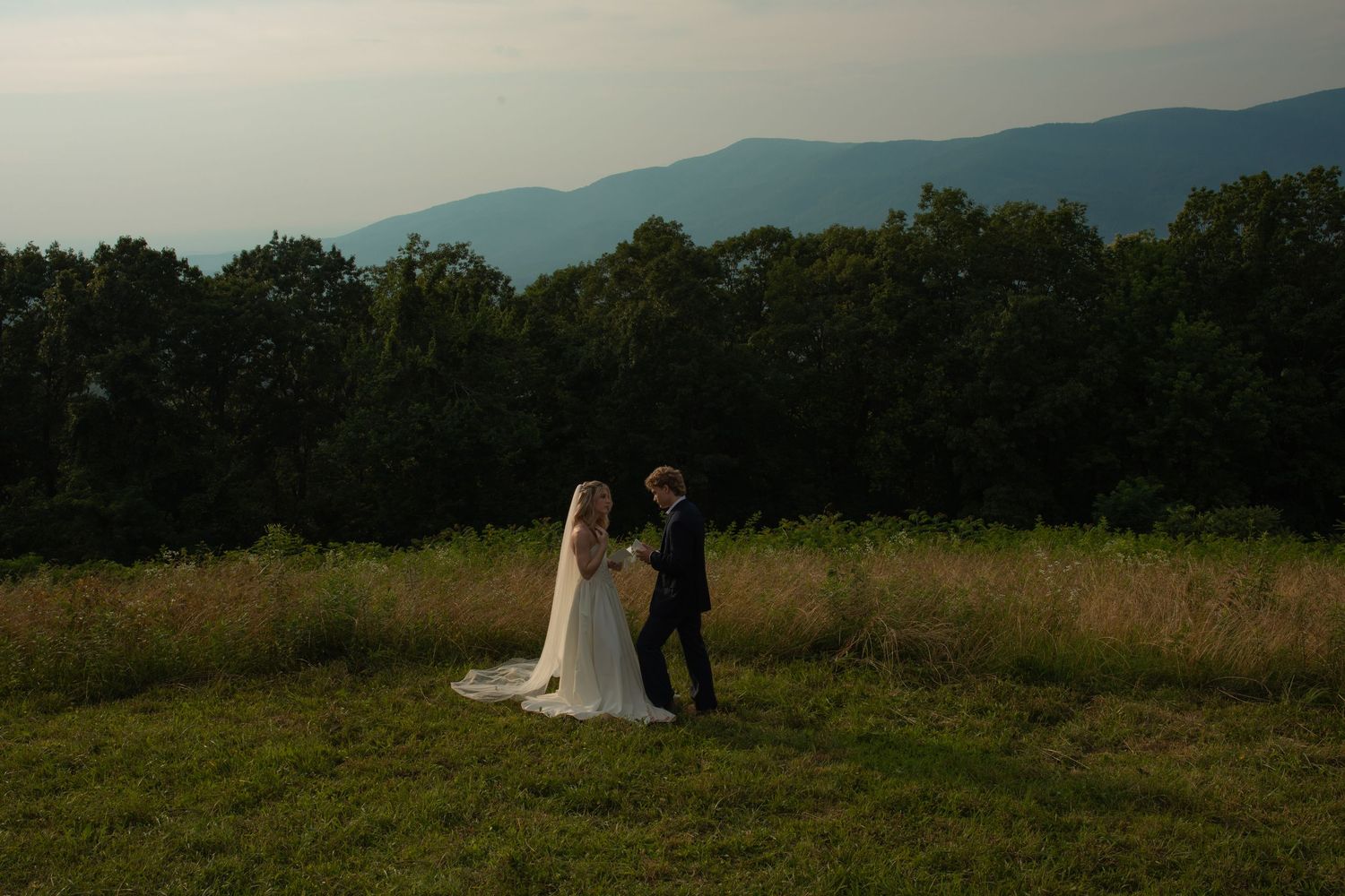 Bride and groom share a romantic moment in a mountain meadow at dusk, with misty blue mountains in the background.