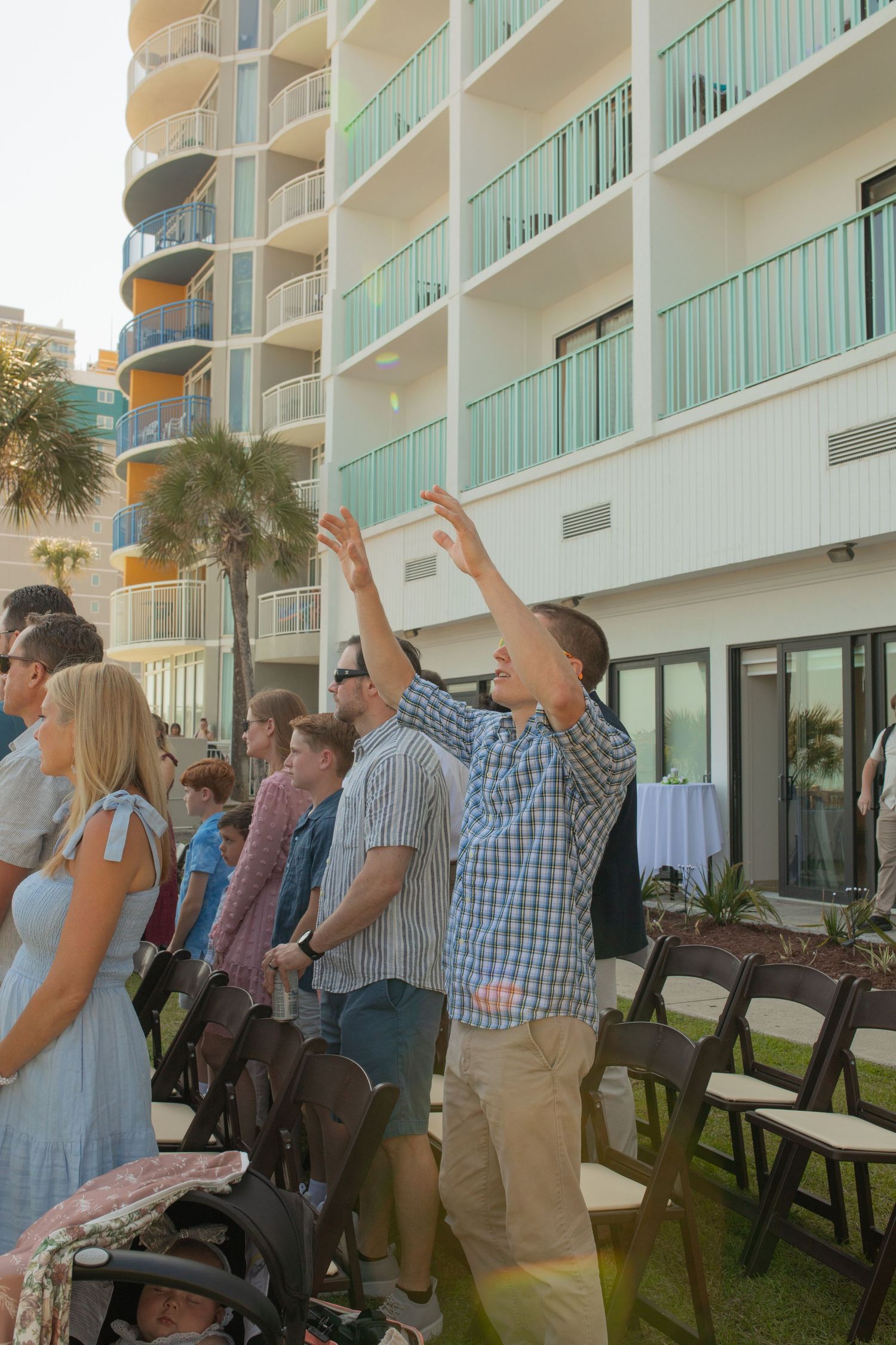 Man raises hands in worship at outdoor gathering near modern beachside hotel with teal balconies and palm trees.