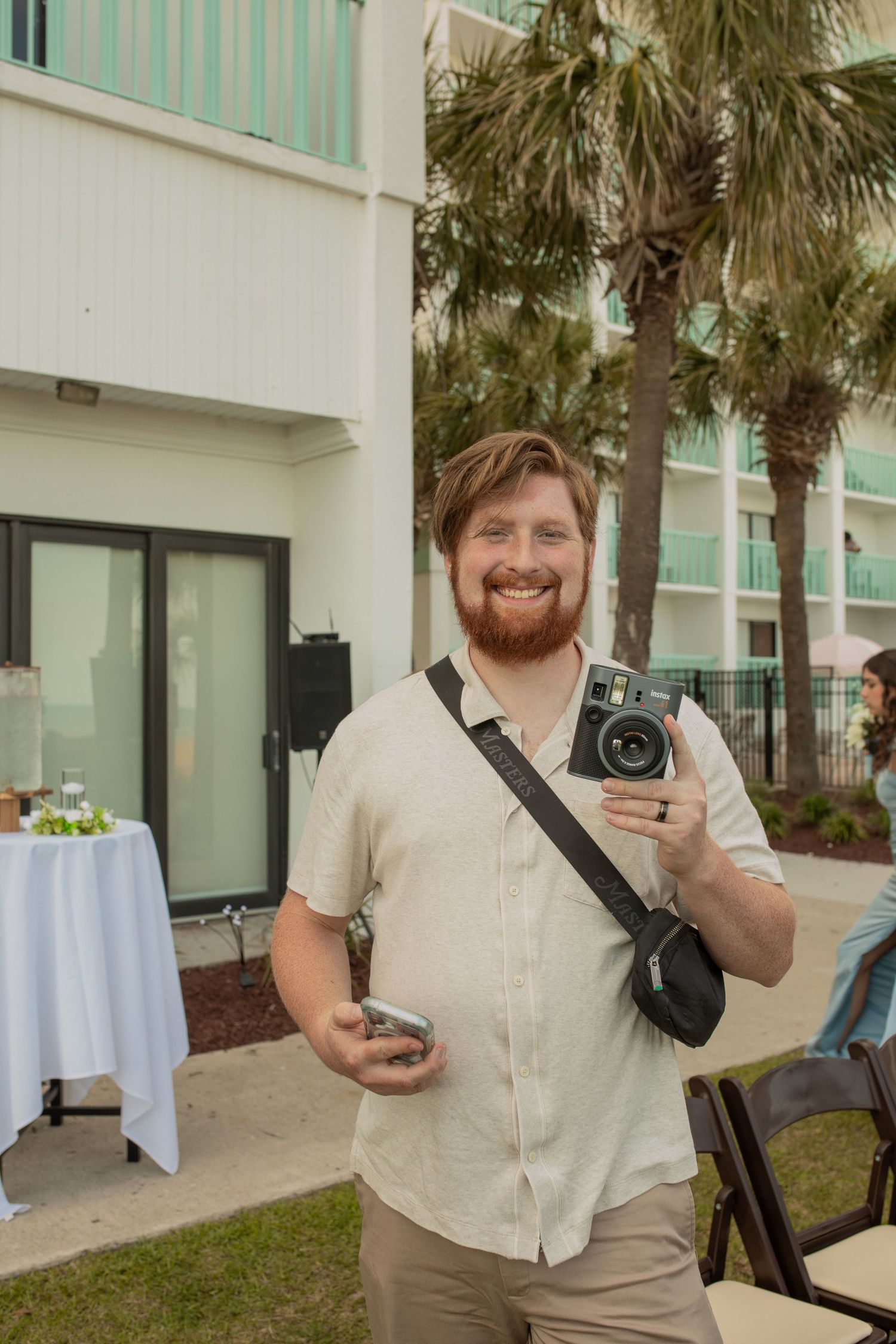 Smiling bearded man holding an instant camera at an outdoor event near palm trees and a teal building.