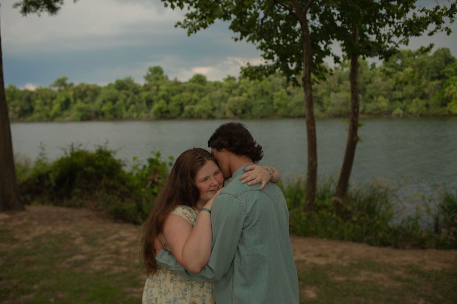 Couple embracing tenderly near a serene river with lush green trees under a cloudy sky.
