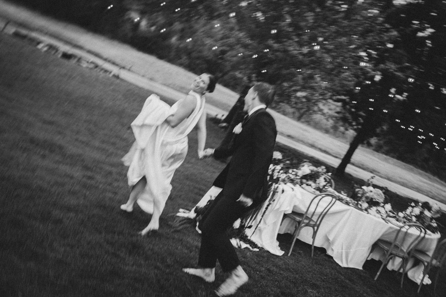 Black and white photo of a bride and groom dancing joyfully outdoors on grass at their wedding reception.