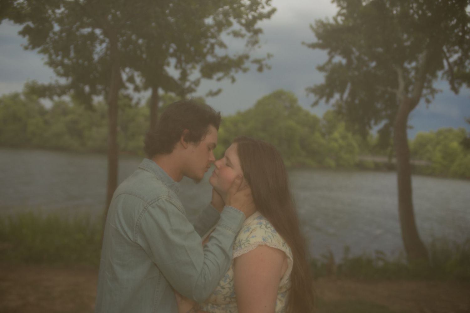 Romantic couple sharing an intimate moment near a serene river surrounded by lush green trees on a cloudy day.