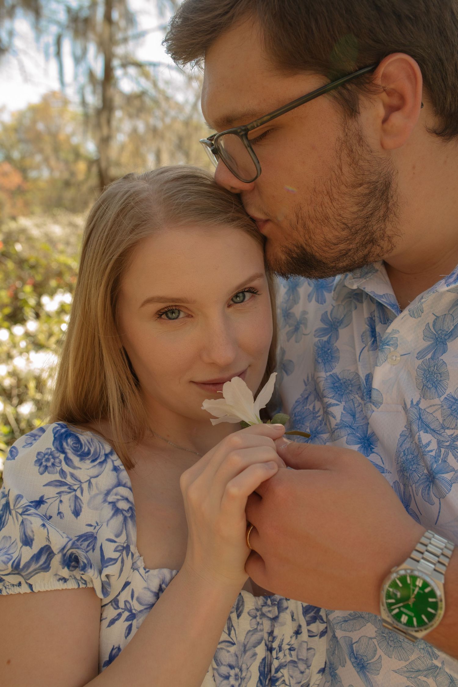 A couple in matching floral outfits shares a tender moment outdoors, as a man kisses a woman's head while she holds a white flower.
