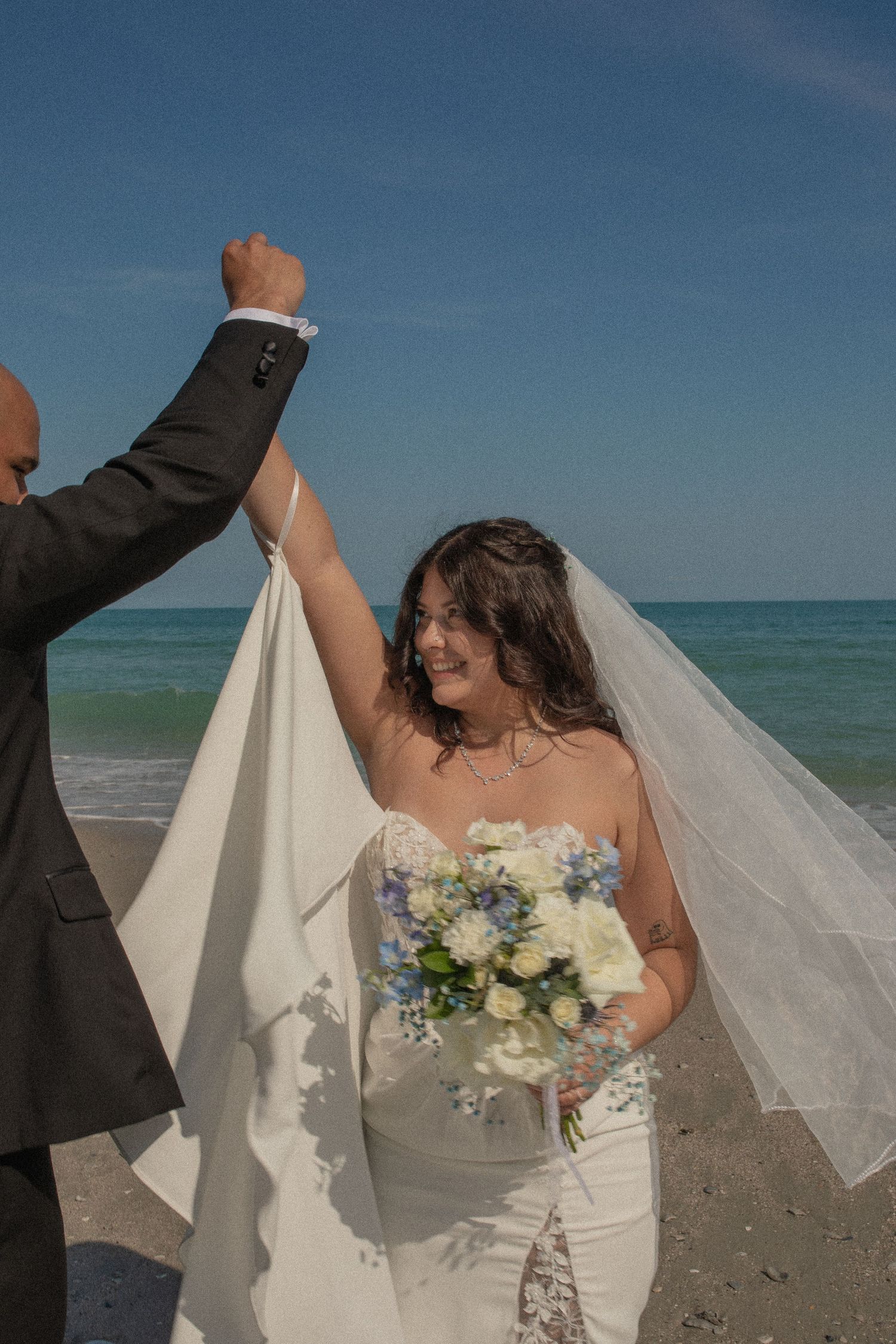 Bride in white strapless gown holds bouquet of blue and white flowers as groom lifts her veil on a sunny beach.