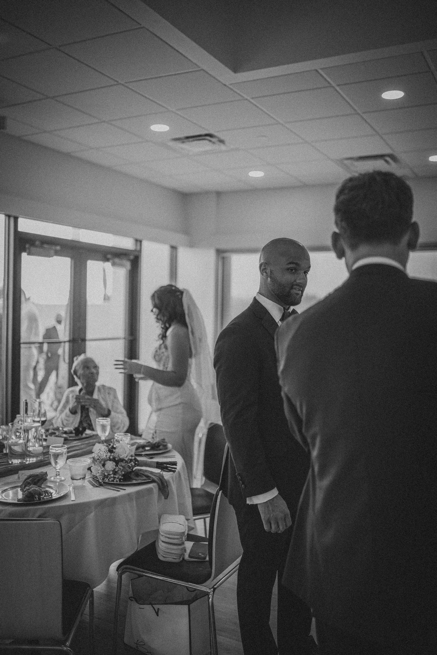 Black and white wedding reception photo showing groom in suit smiling as bride in veil stands near elegantly set dining tables.