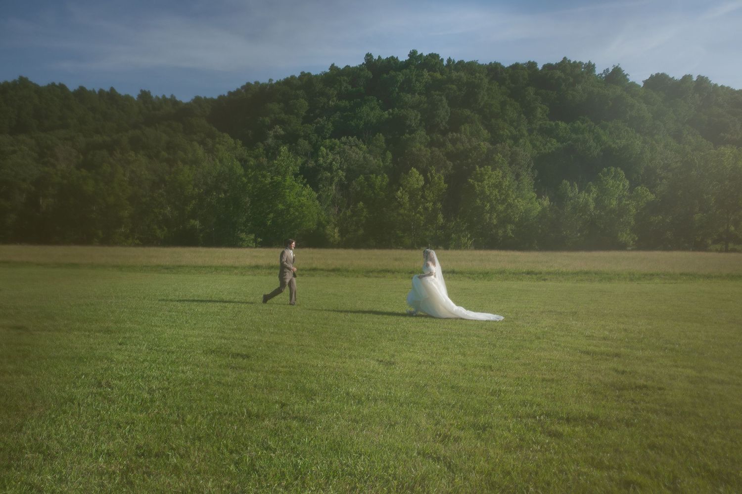 A bride in a flowing white gown and groom run across a lush green field with a forested hill backdrop.