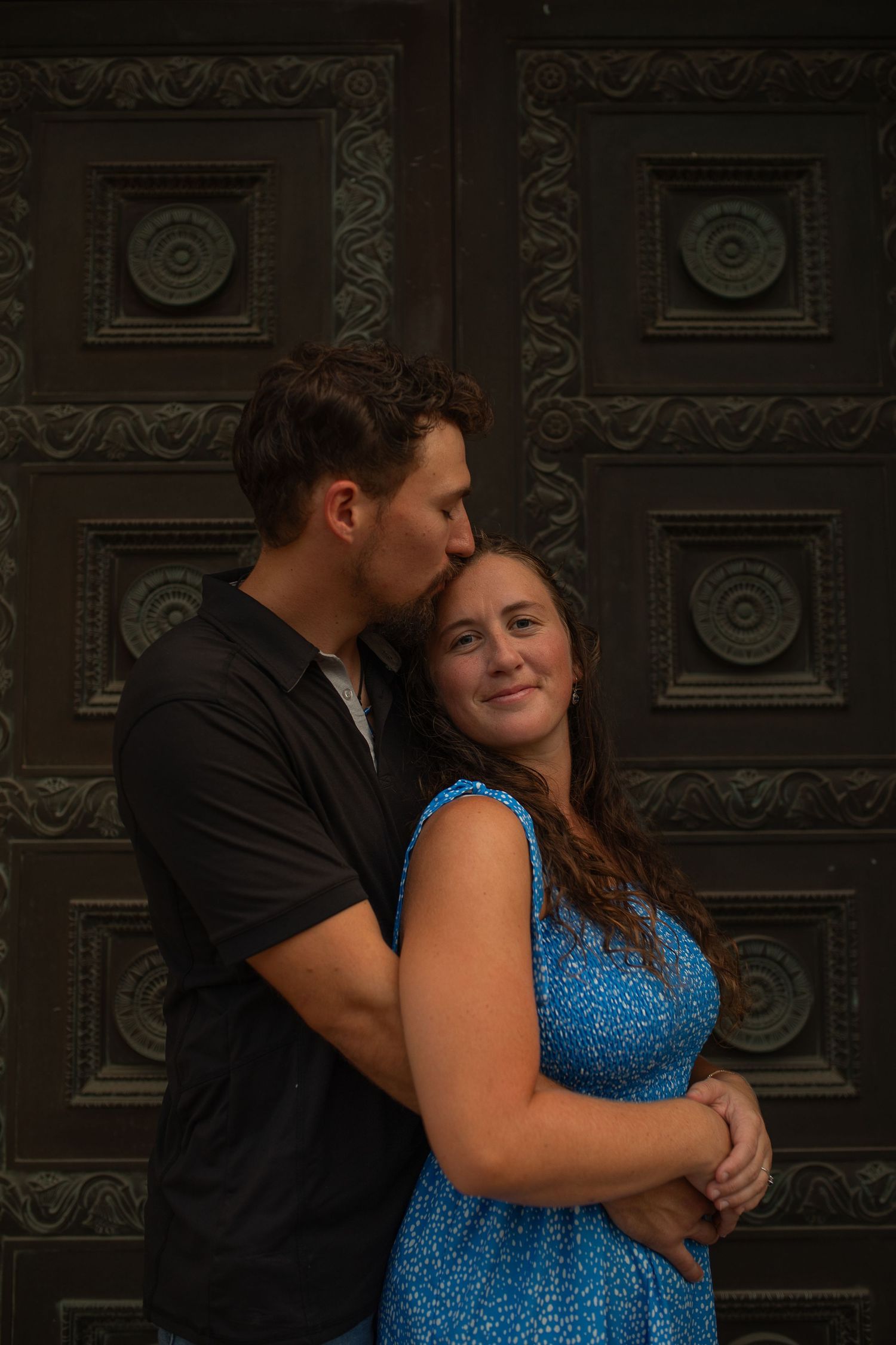 A couple poses romantically before an ornate dark door, man kissing woman's temple as she smiles in a blue floral dress.