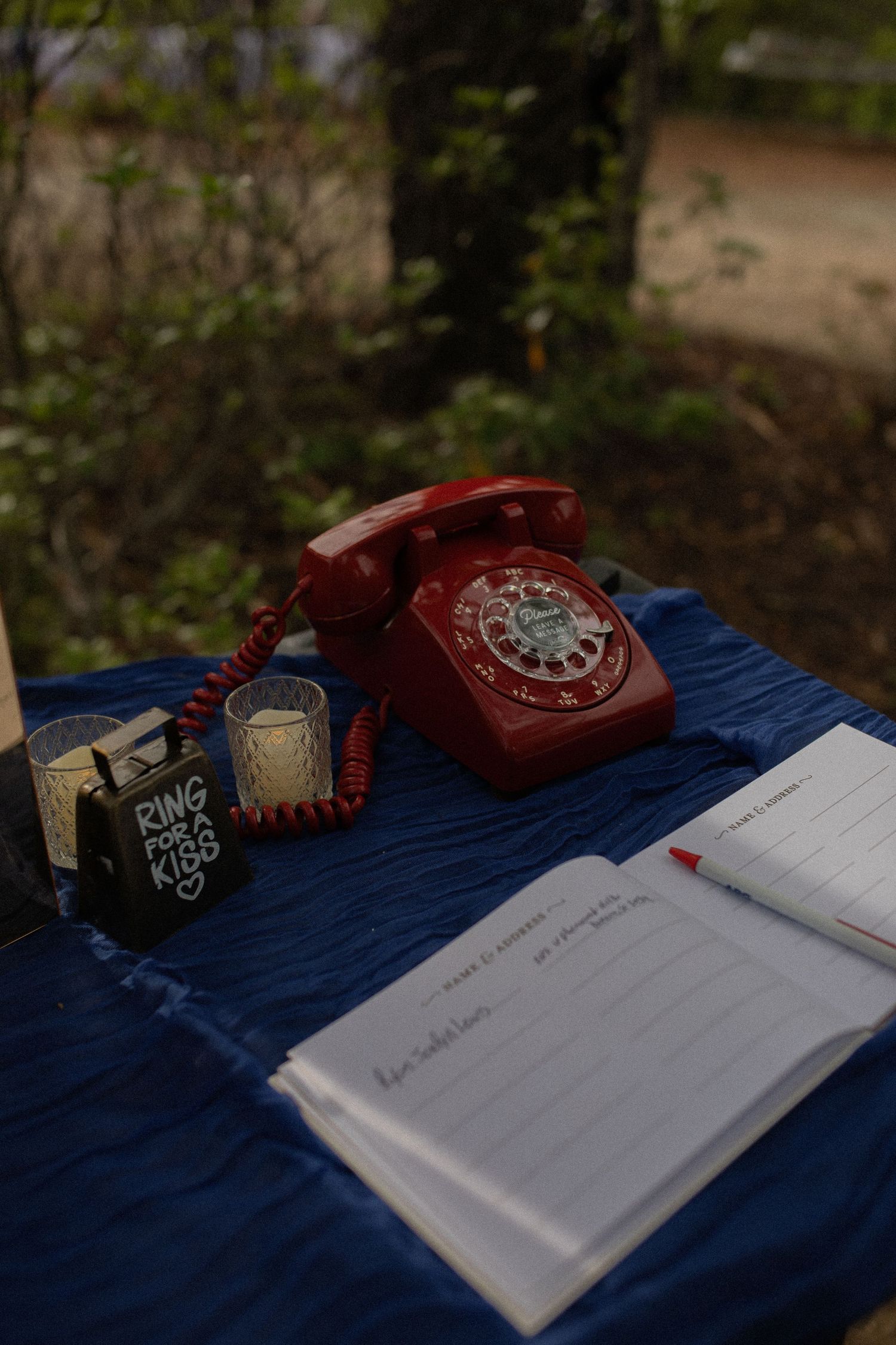 Red rotary phone beside open guest book and candles on blue tablecloth at outdoor wedding reception.