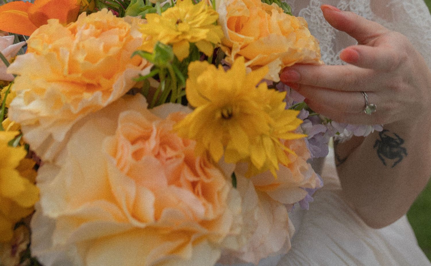 Bride holding a vibrant wedding bouquet of peach roses and yellow daisies, wearing a diamond ring and floral tattoo.