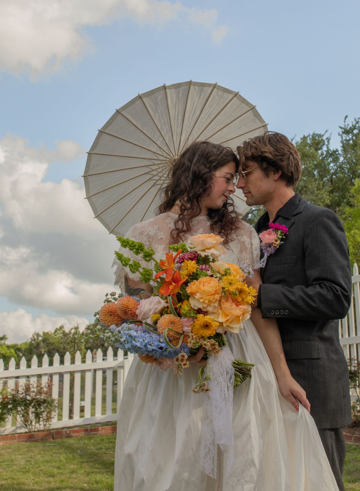 Bride and groom share romantic moment under white parasol, bride holding colorful bouquet, outdoors near white picket fence.