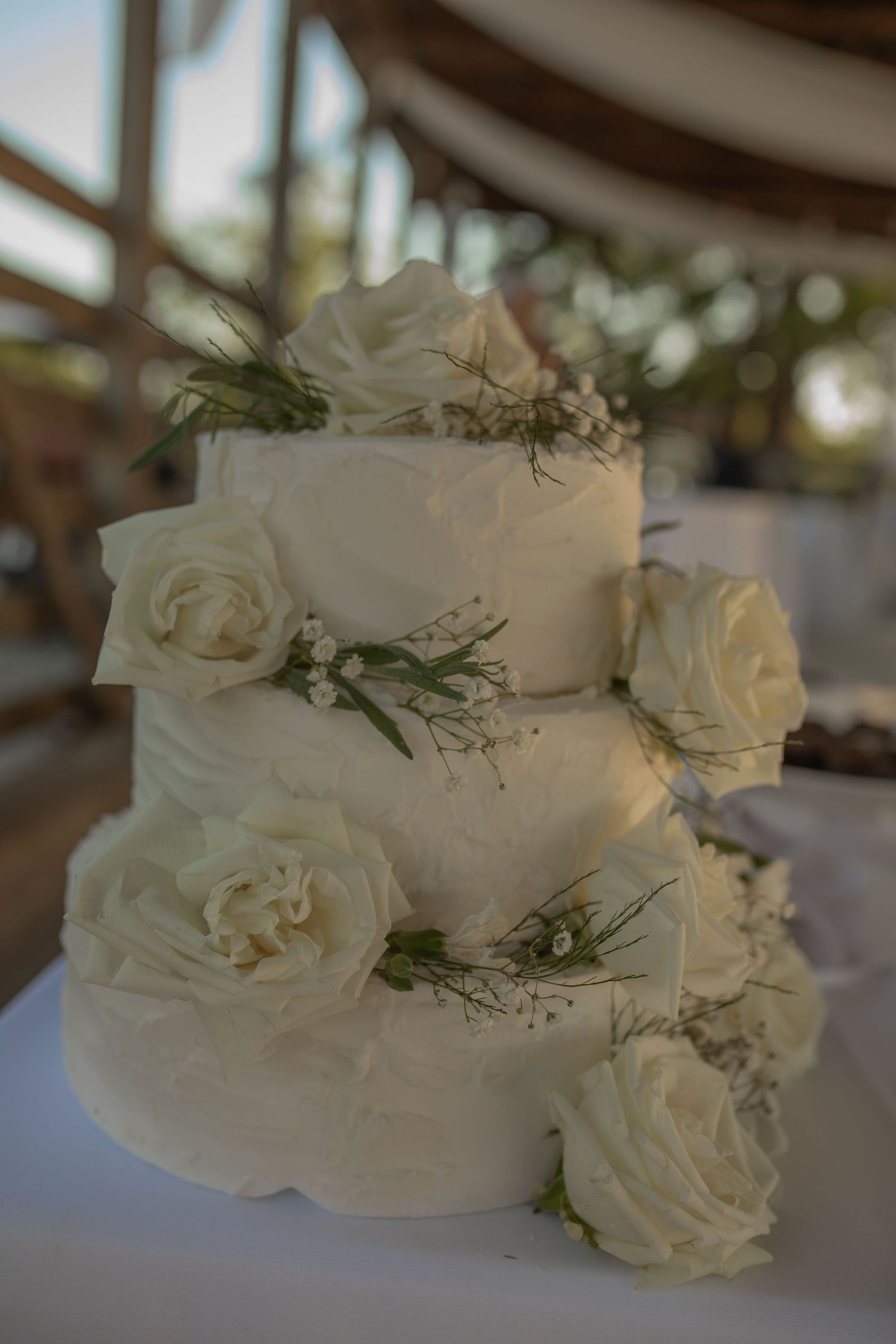 Three-tier white wedding cake decorated with fresh white roses and greenery on a white tablecloth.