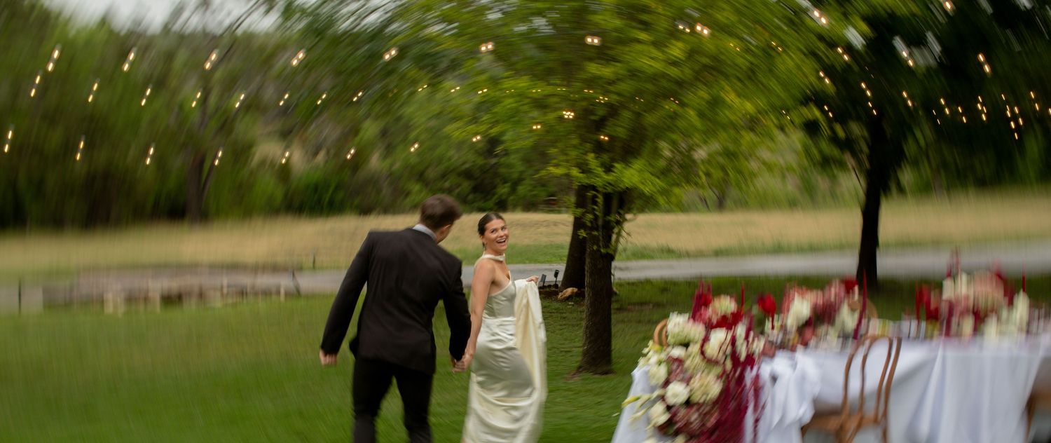 A bride and groom walk hand in hand at an outdoor wedding reception with string lights and floral table settings.