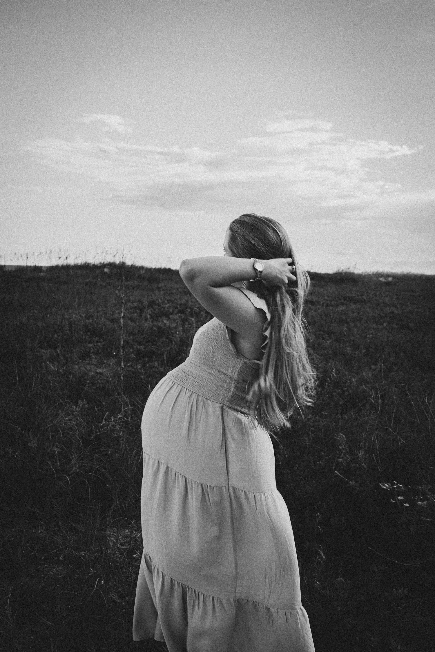 A flowing dress silhouette stands in a field against cloudy sky in black and white.