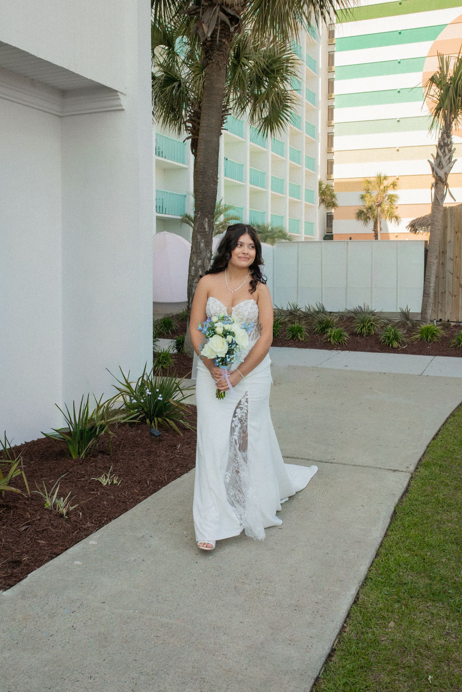 Bride in white lace gown holding bouquet stands on pathway outside hotel with palm trees.