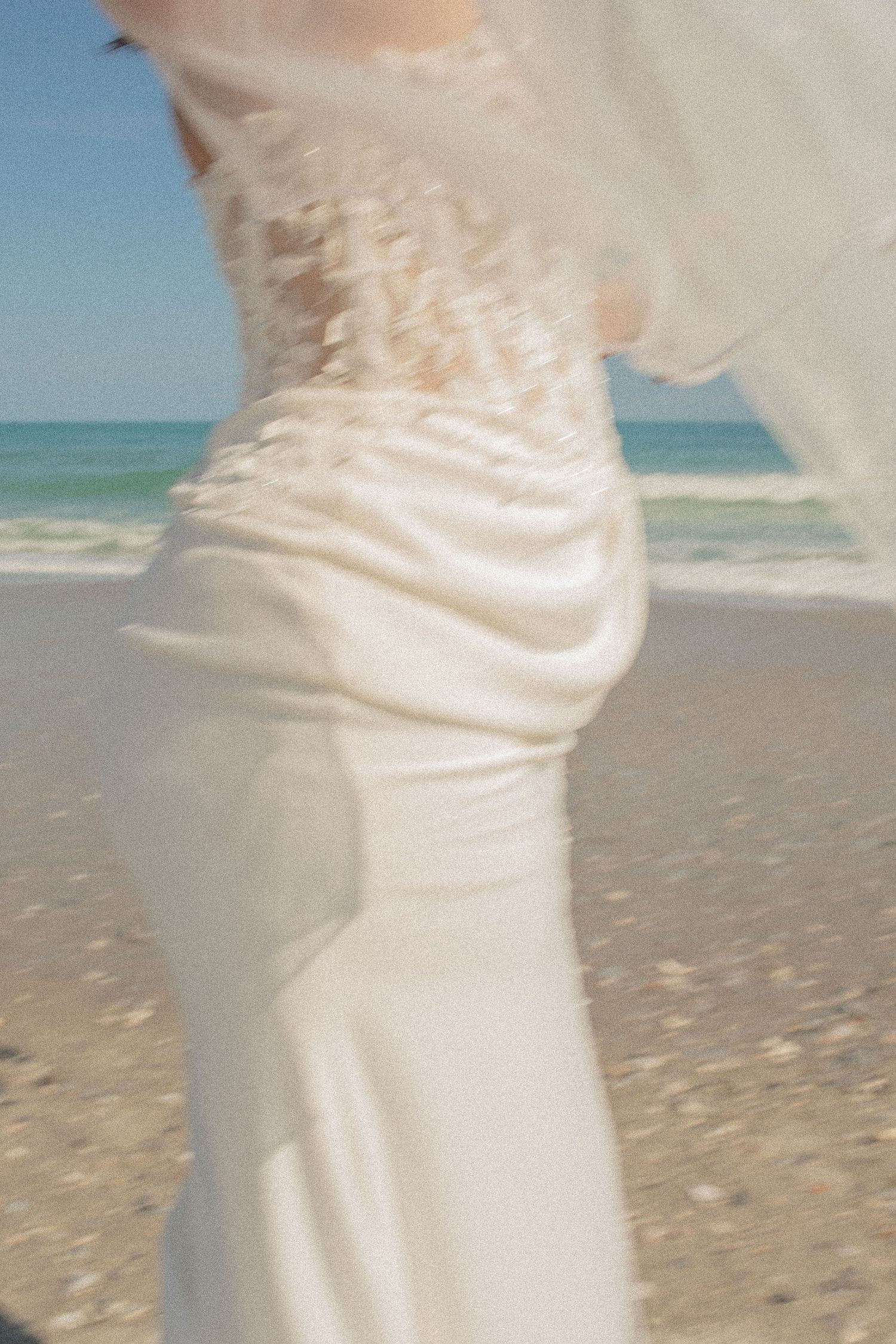 A bride in a flowing white wedding gown and veil stands on a sandy beach with ocean waves in the background.